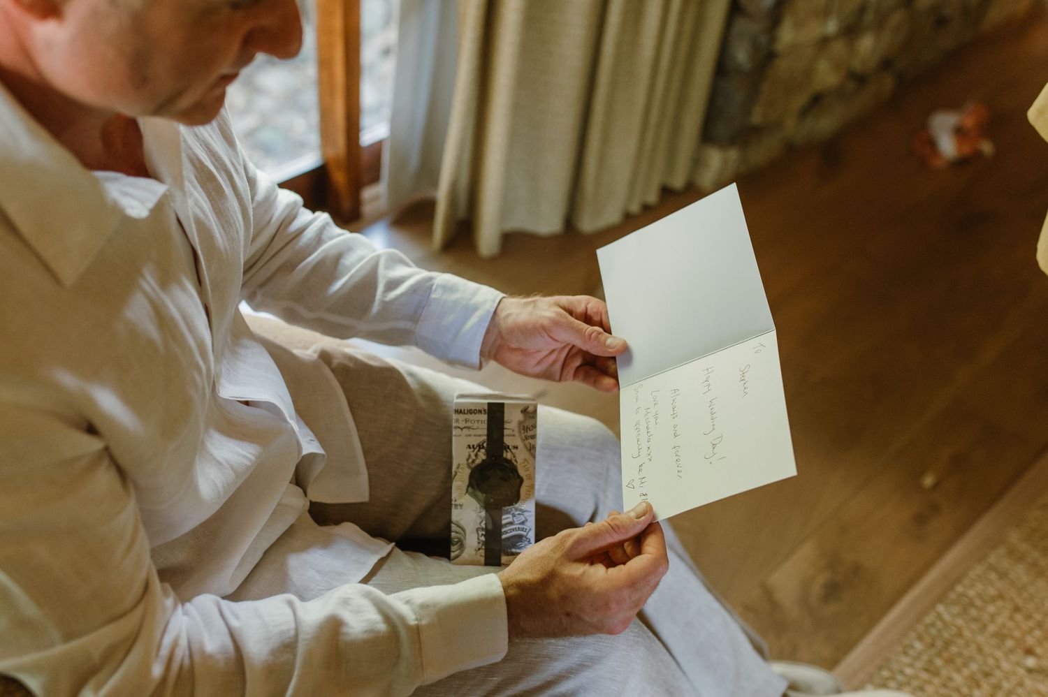 Man in white shirt reading a greeting card with a gift bottle indoors.
