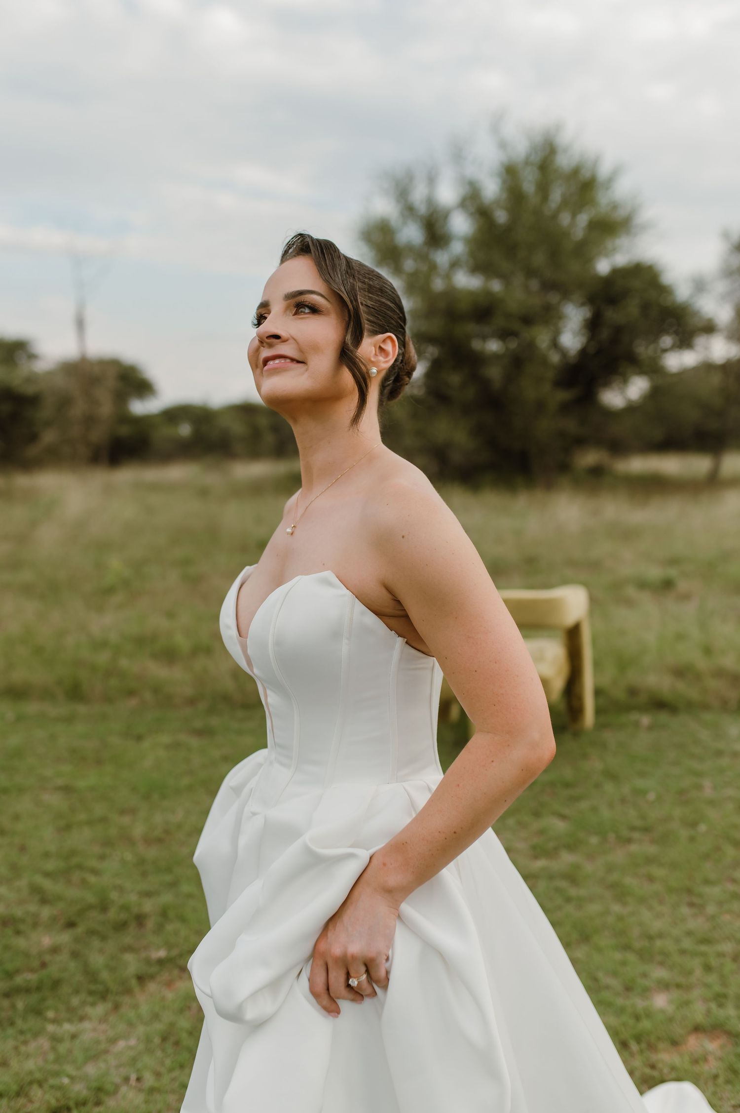 A bride in a white strapless wedding gown poses in a sunlit meadow with trees in the background.