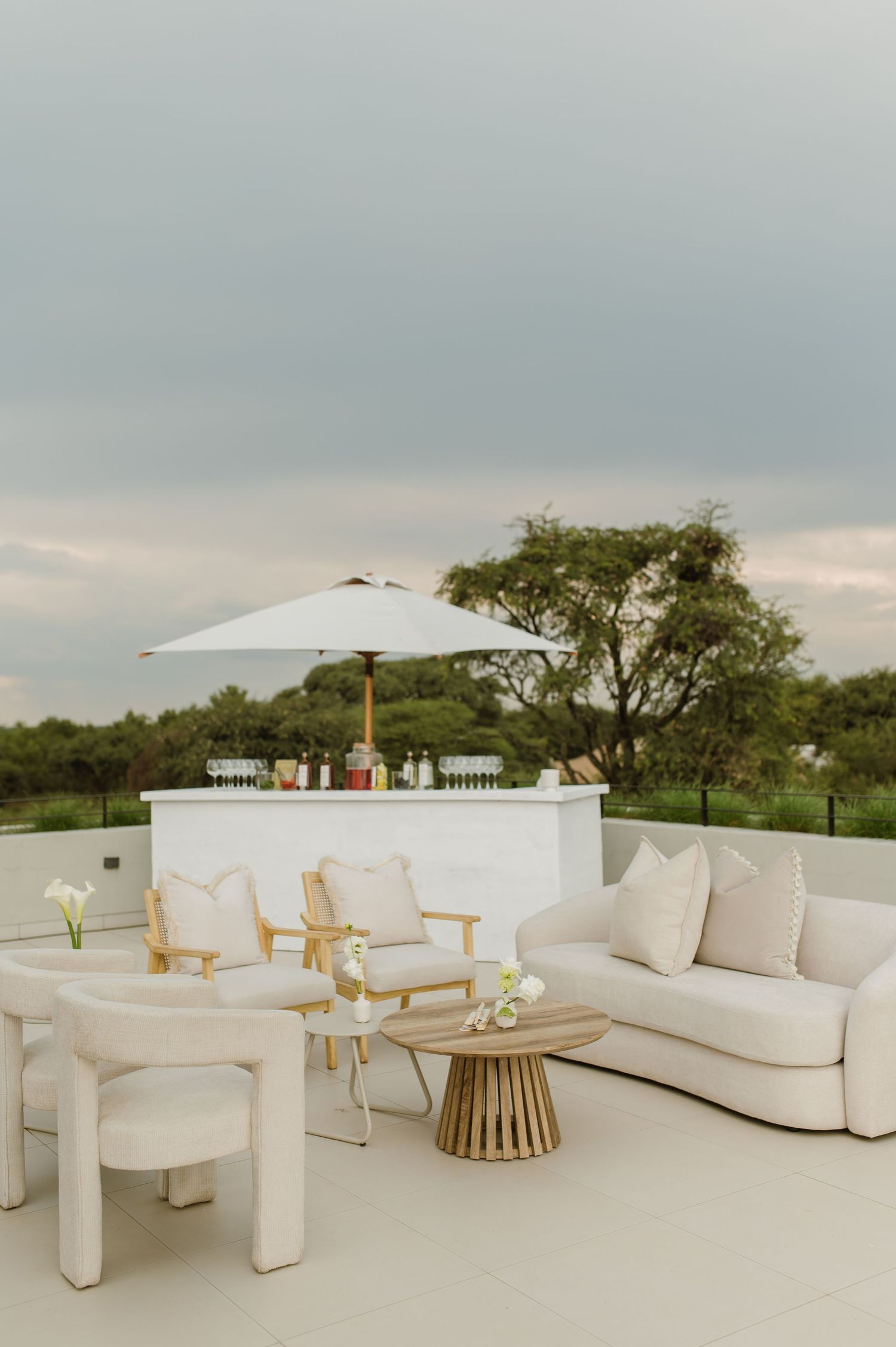 Elegant outdoor lounge area with white furniture, umbrella and bar setup on a rooftop terrace overlooking nature.