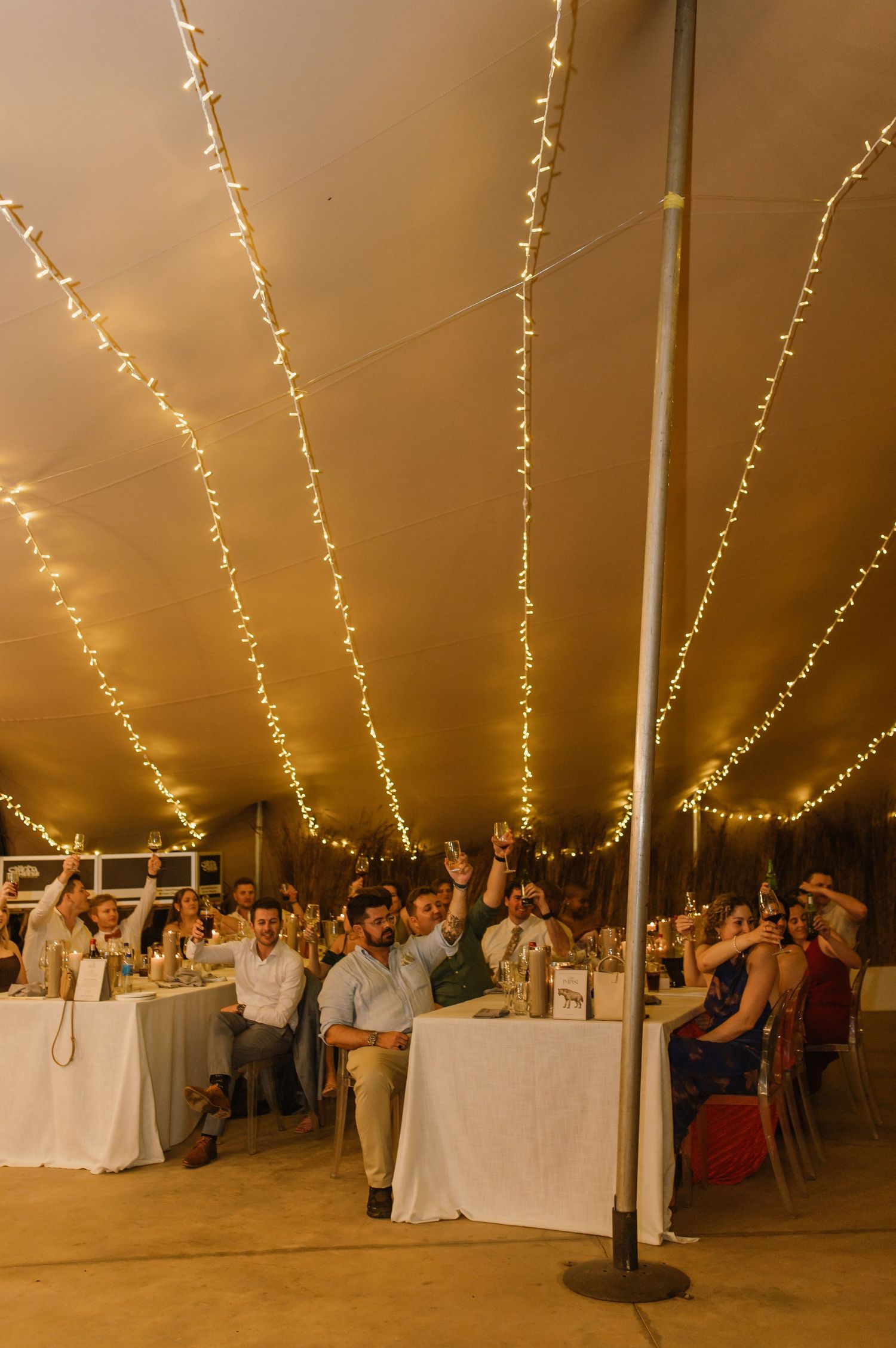 Guests celebrating at a tented reception with string lights and white-clothed tables.
