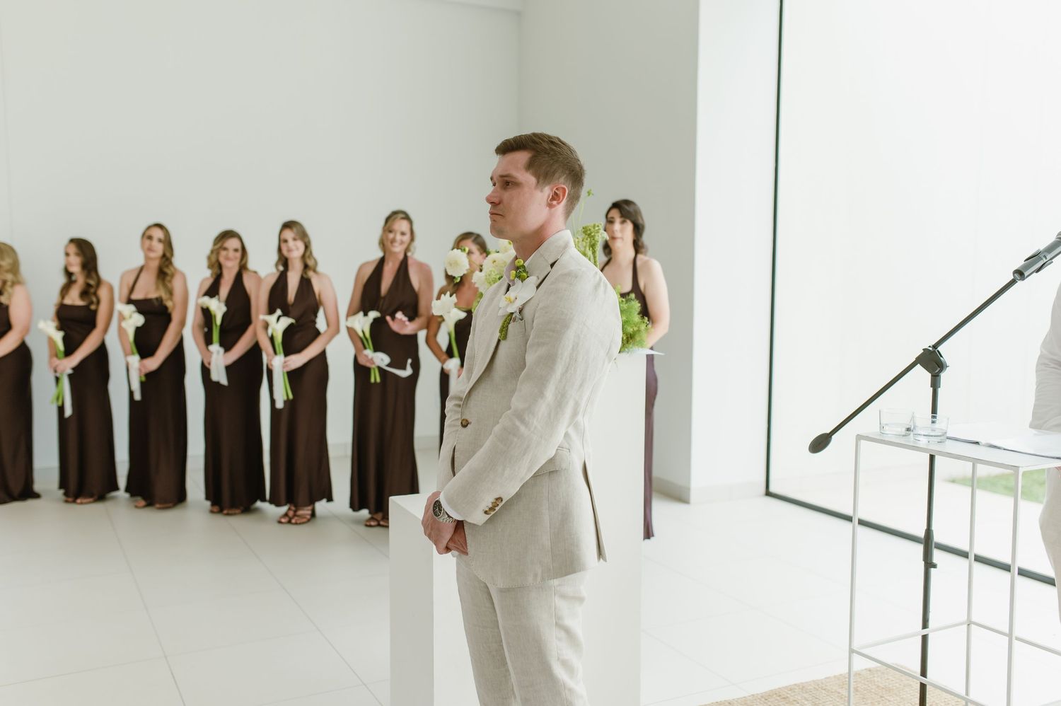 A wedding ceremony with bridesmaids in black dresses standing in line against a white wall.