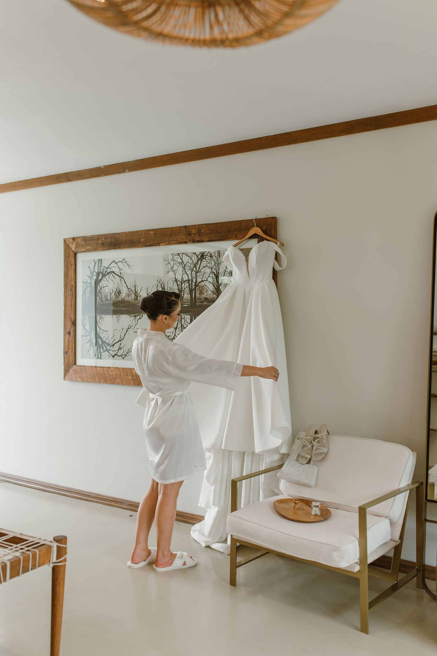A white wedding dress hangs elegantly on display in a minimalist room with natural lighting.
