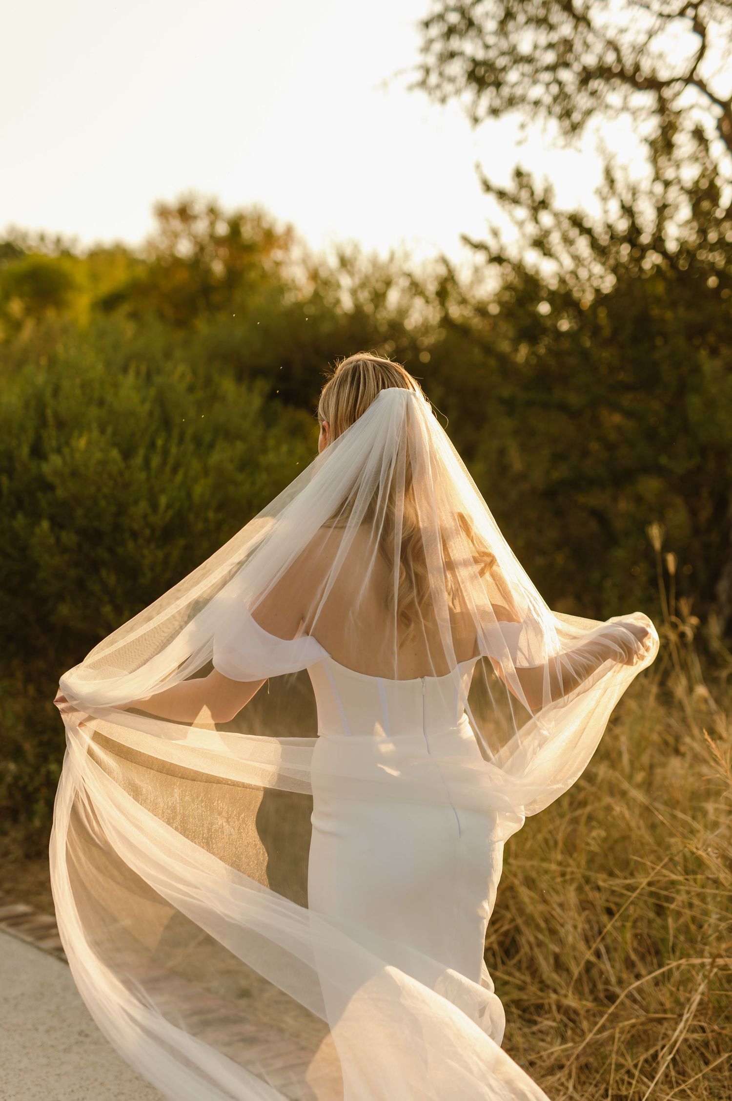 Bride plays with flowing veil creating ethereal silhouette against sunset light.