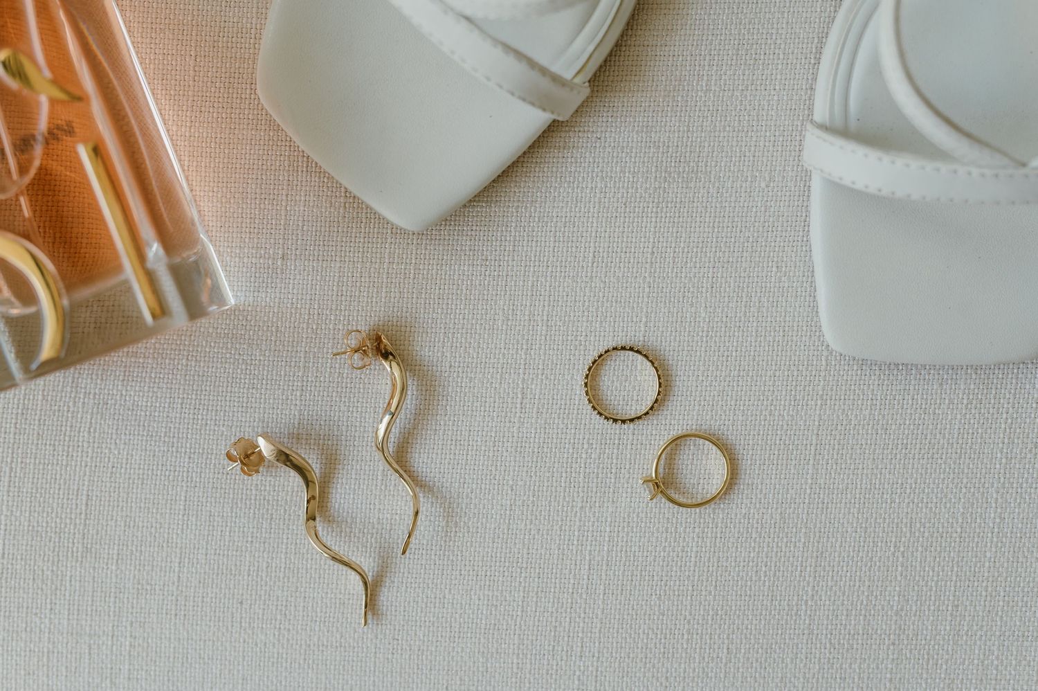Wedding accessories including white shoes and gold jewelry on neutral surface.