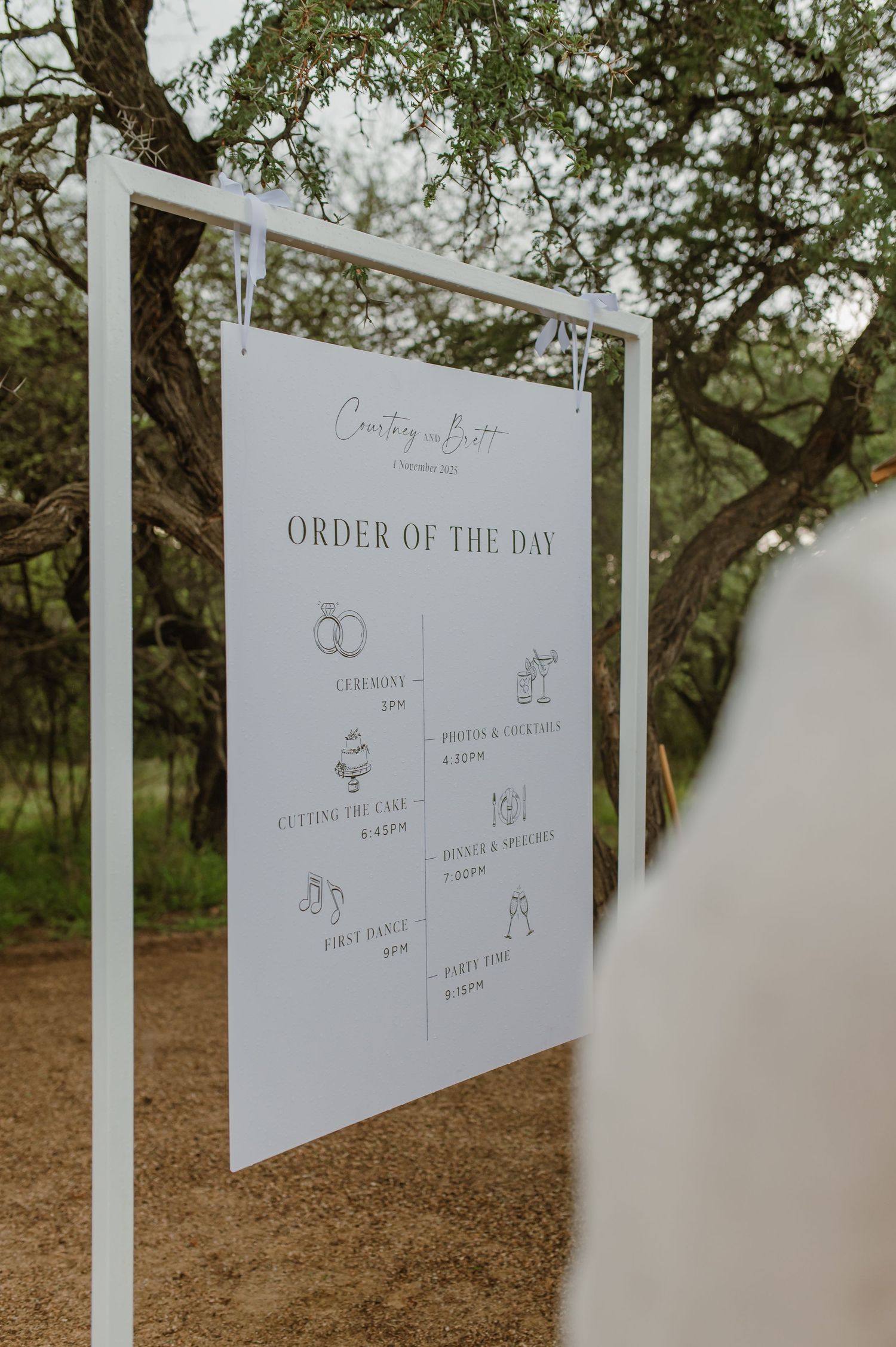 White wedding schedule sign displaying order of the day mounted on poles outdoors.