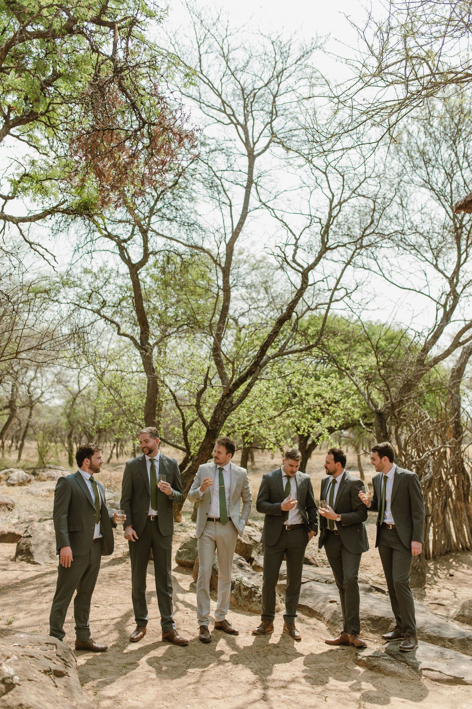Groomsmen in suits standing in a line among trees in a rustic outdoor setting.