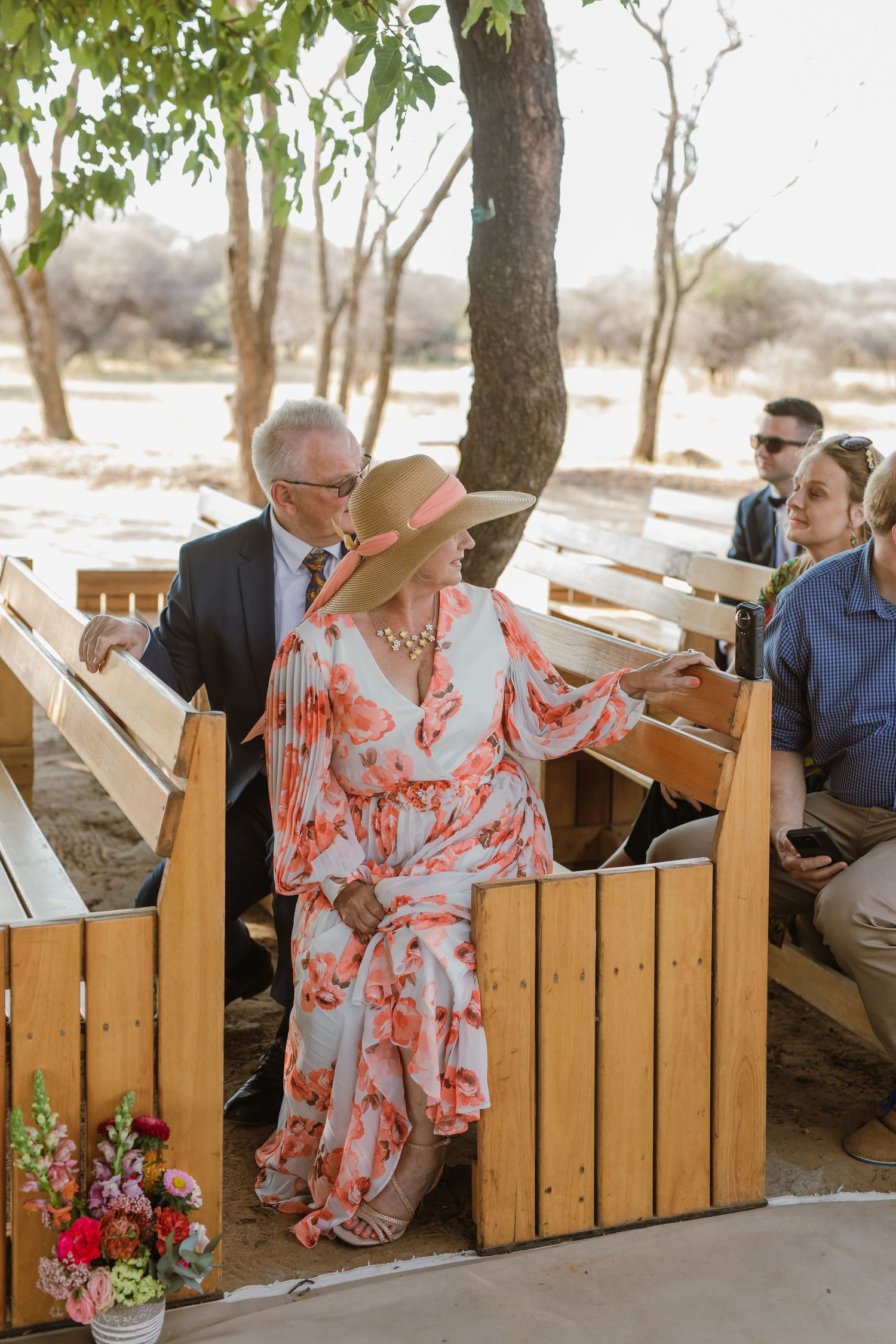 Guests seated on wooden benches at an outdoor wedding venue under leafy trees.