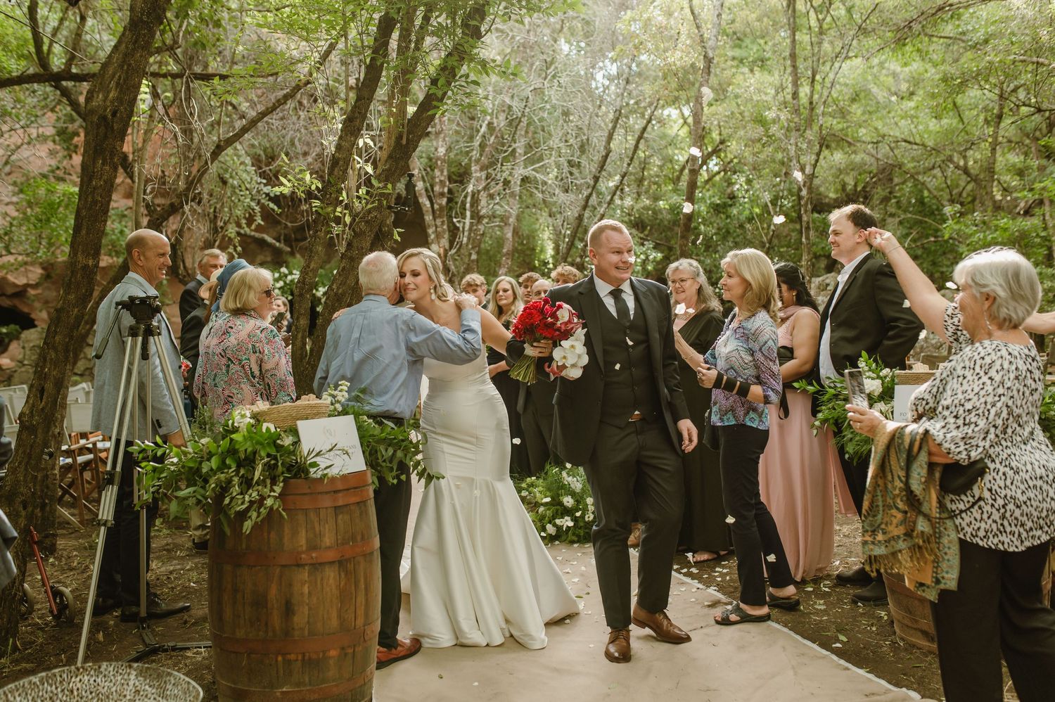Outdoor wedding celebration with guests mingling on a tree-lined path in dappled sunlight.