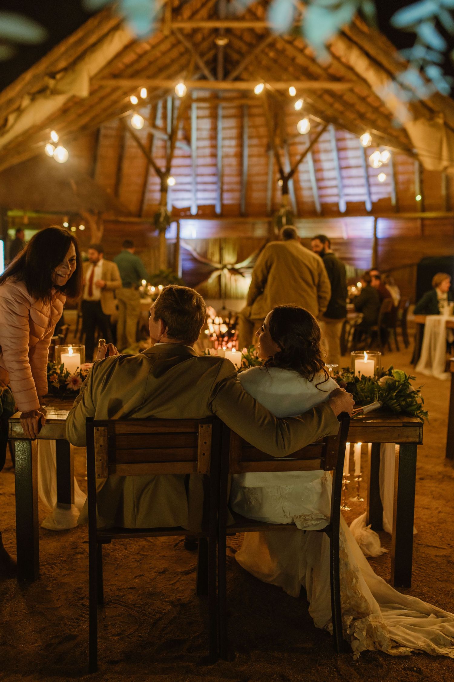 Intimate candlelit seating area with wooden benches under a rustic barn ceiling with string lights.