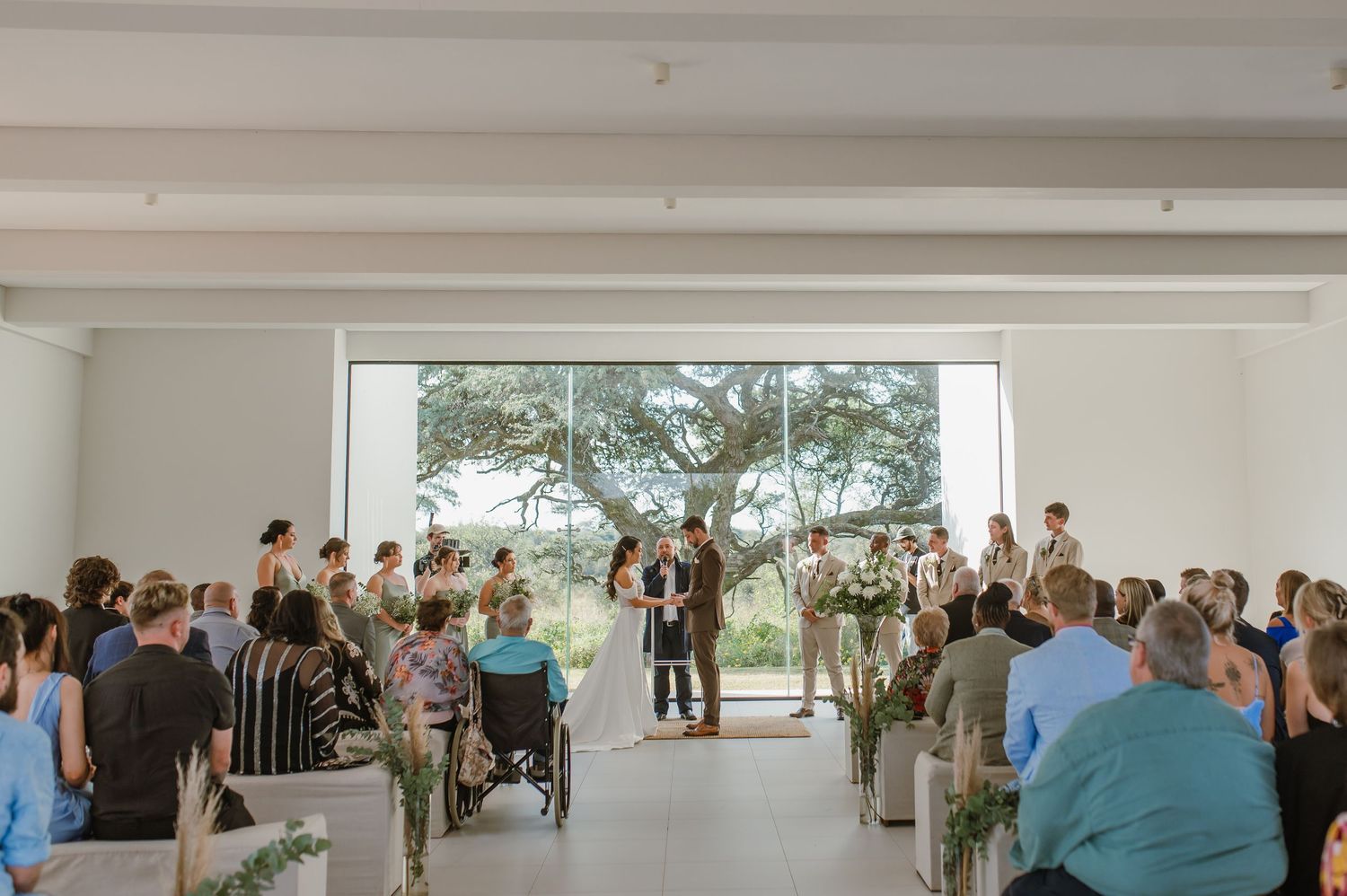 Wedding guests gather in a modern minimalist venue with white walls and large windows.