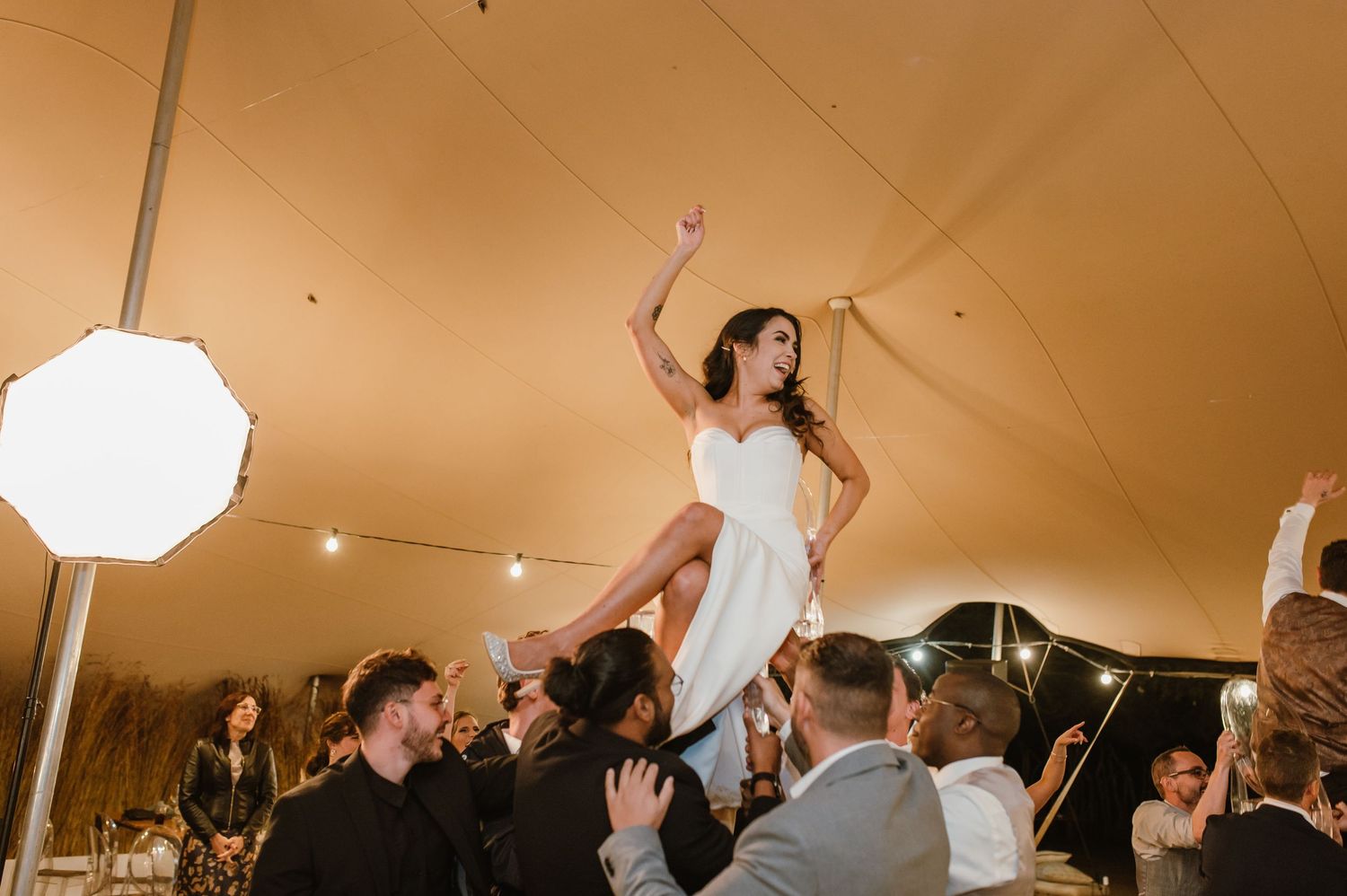 A person in a white dress is lifted high by guests during an energetic wedding reception celebration.