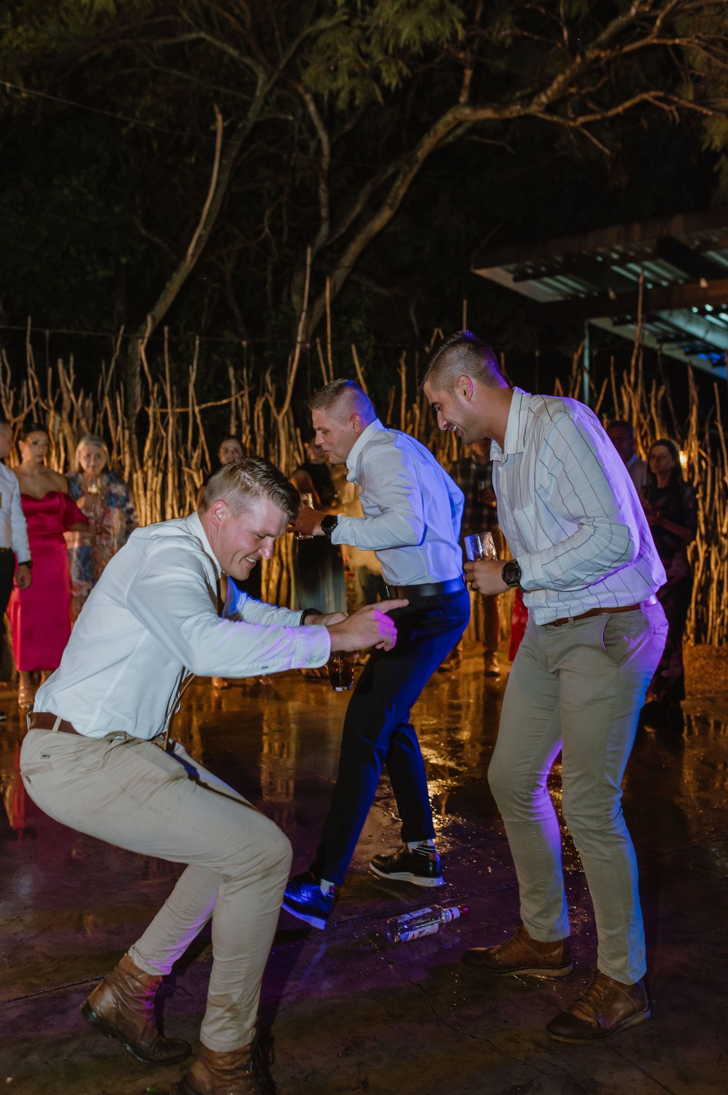 Three men dancing enthusiastically on outdoor dance floor at nighttime wedding reception party.