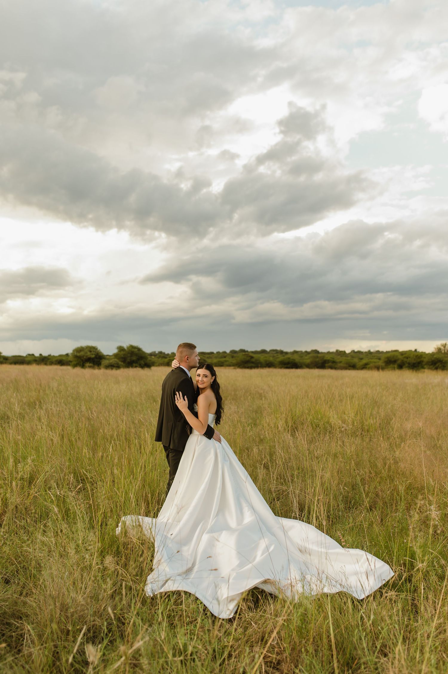 Bride in flowing white gown with train embraces groom in dramatic open field under cloudy sky.