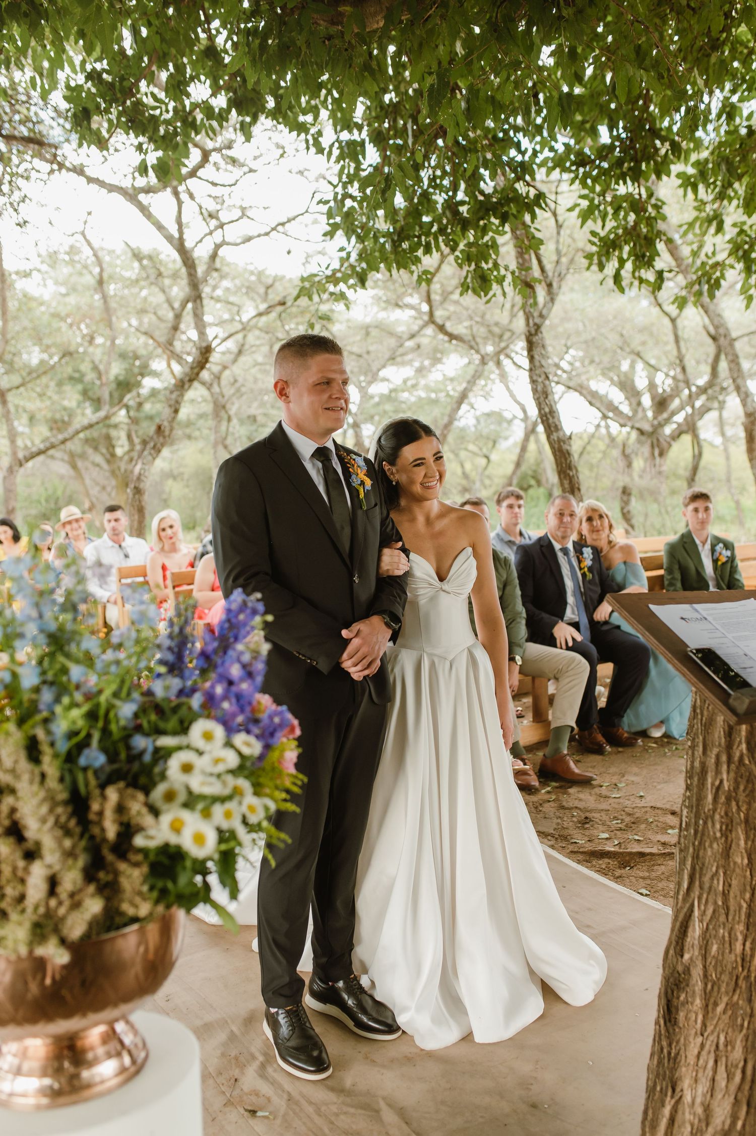 Bride and groom stand arm in arm at outdoor ceremony beneath lush green trees.