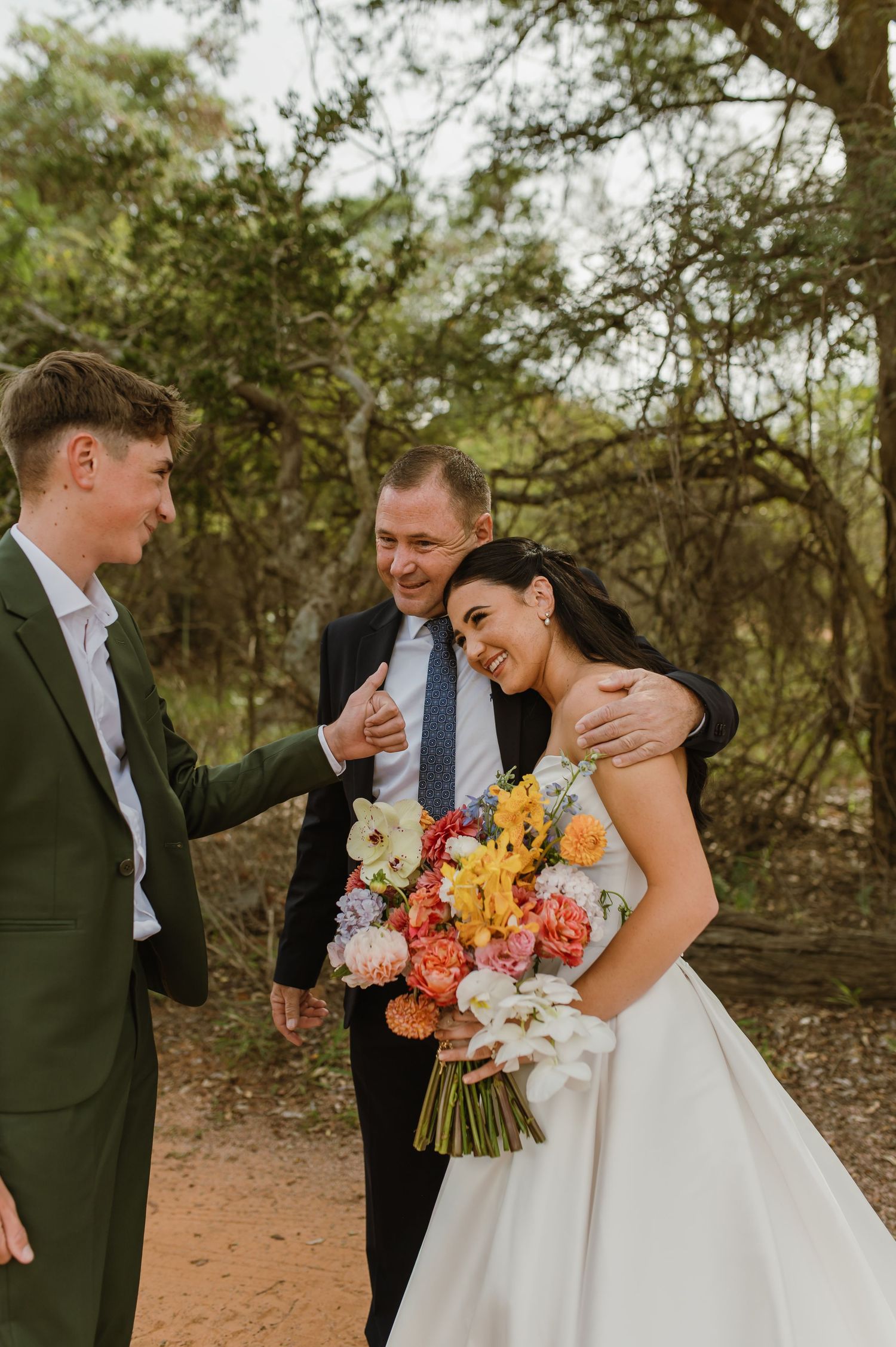 Bride laughing with groom and groomsman outdoors holding vibrant floral bouquet in wooded setting.