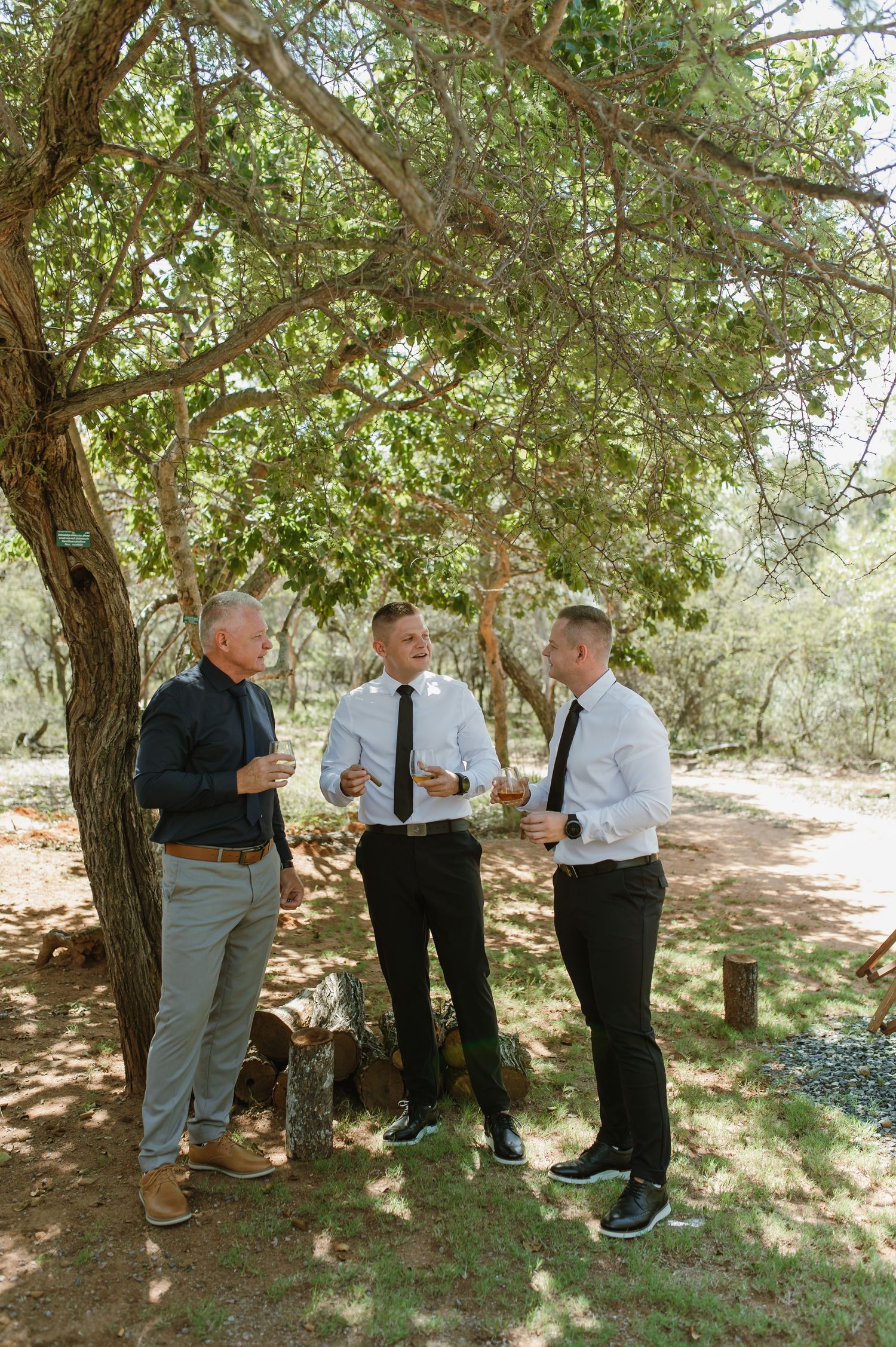 Three men in formal attire socializing under a tree at an outdoor wedding celebration.