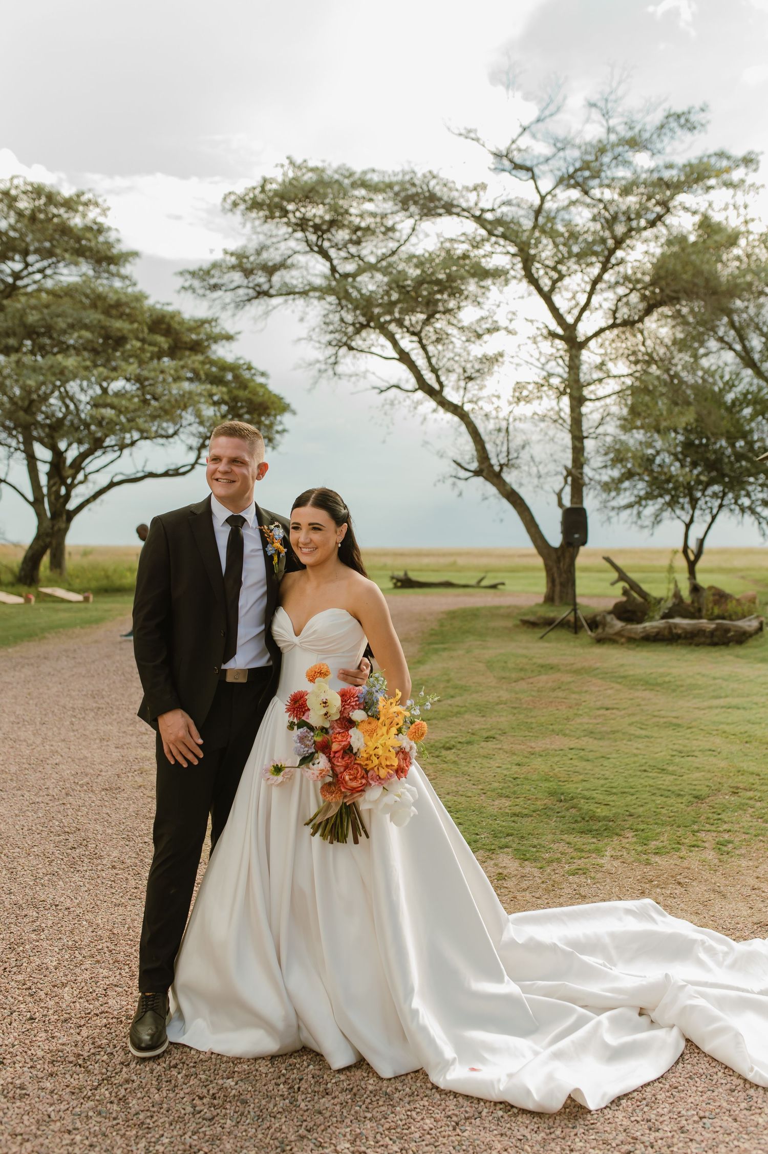 Bride and groom pose on a gravel path with trees and open landscape behind them on their wedding day.