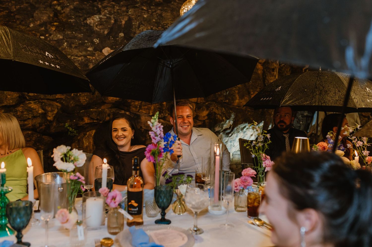 Wedding guests smile under black umbrellas at candlelit reception table with colorful flowers.