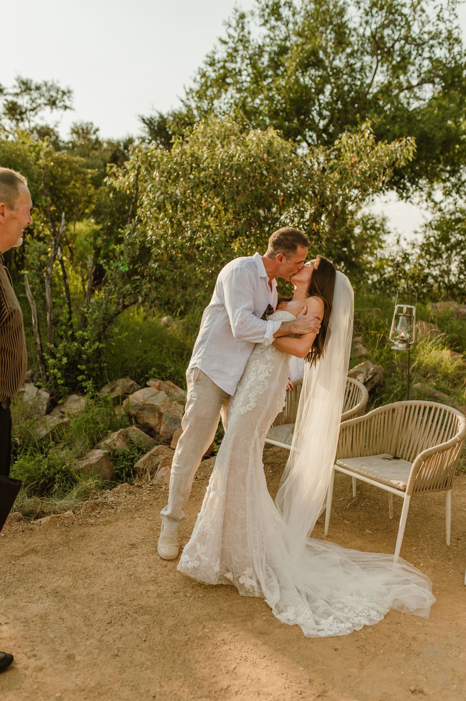 Bride and groom share a first kiss in a romantic outdoor wedding ceremony surrounded by lush greenery.