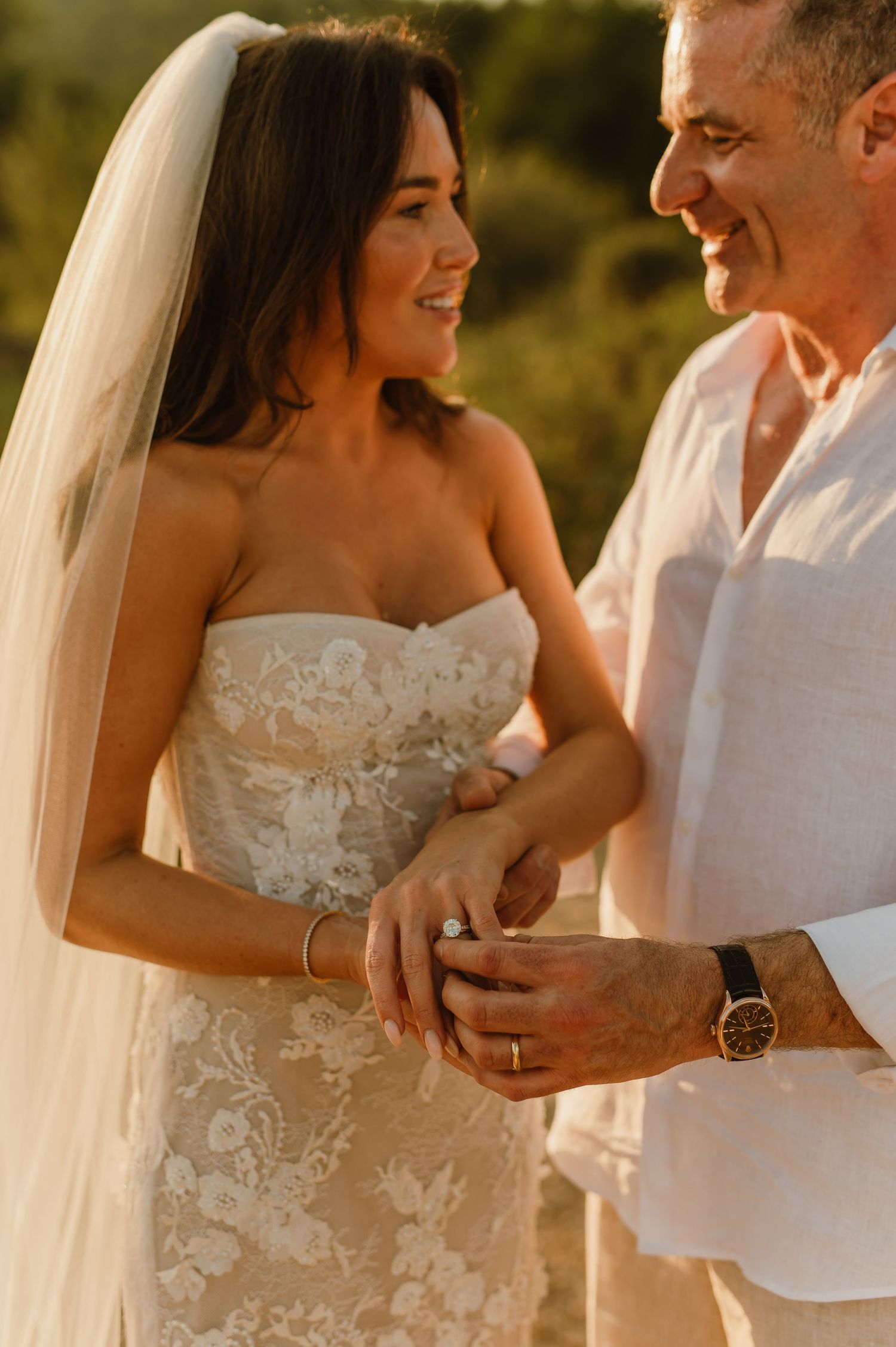 Smiling newlyweds hold hands showing wedding ring, bride in floral lace gown and veil at sunset.