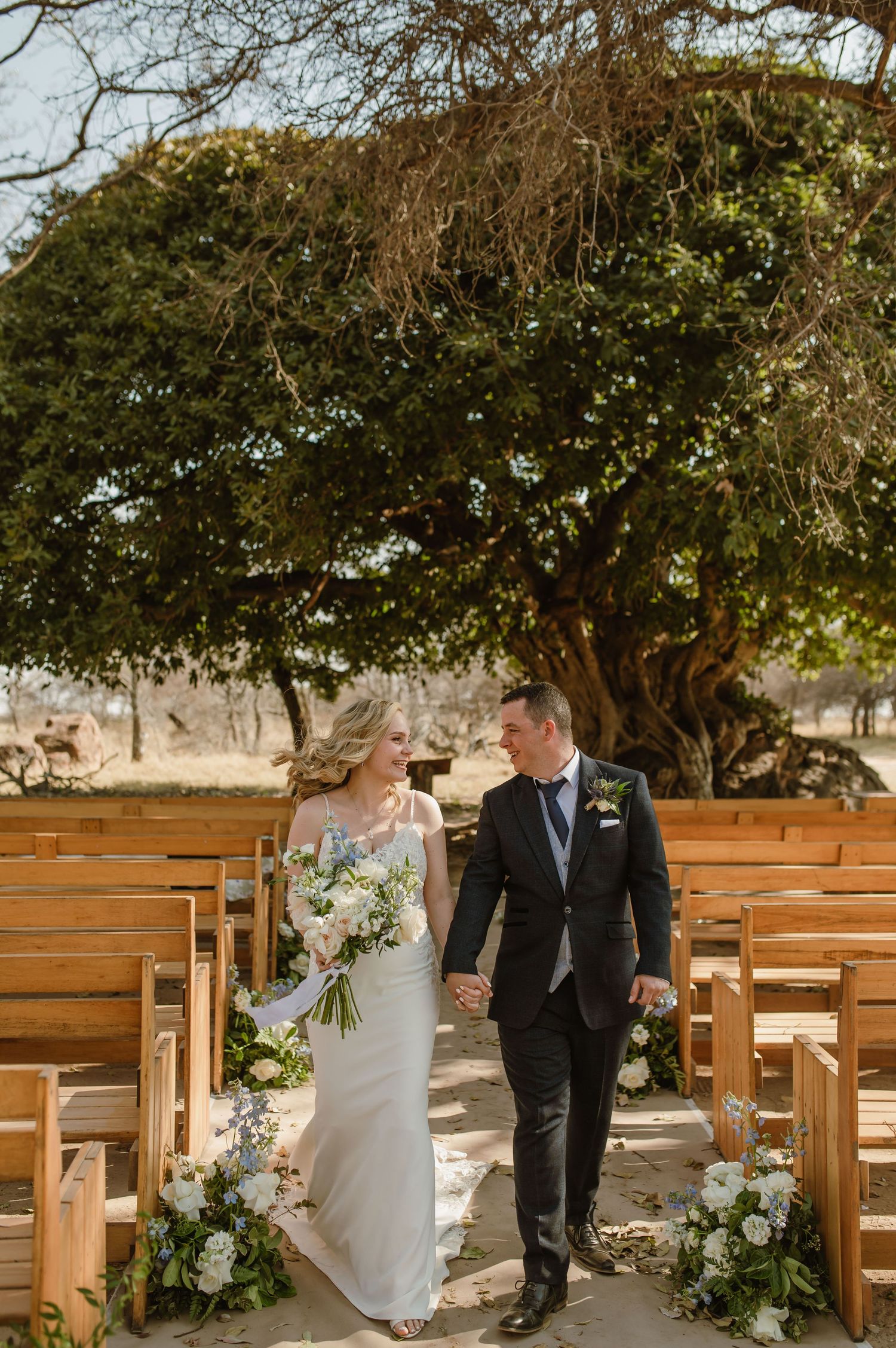 Outdoor wedding ceremony setup with wooden benches and floral decorations under large tree.