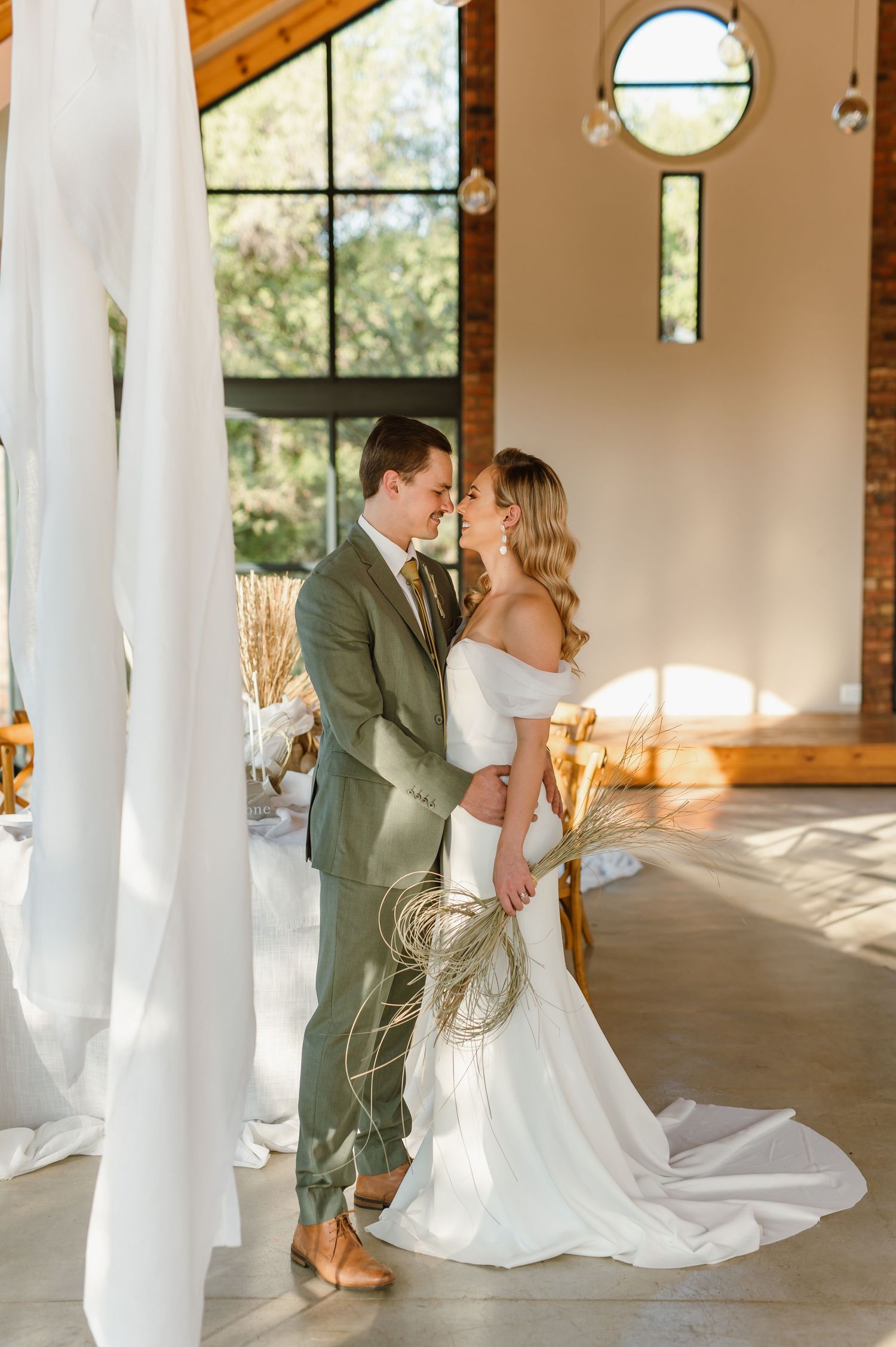 A wedding couple shares an intimate moment in a sunlit venue with large windows and white drapes.