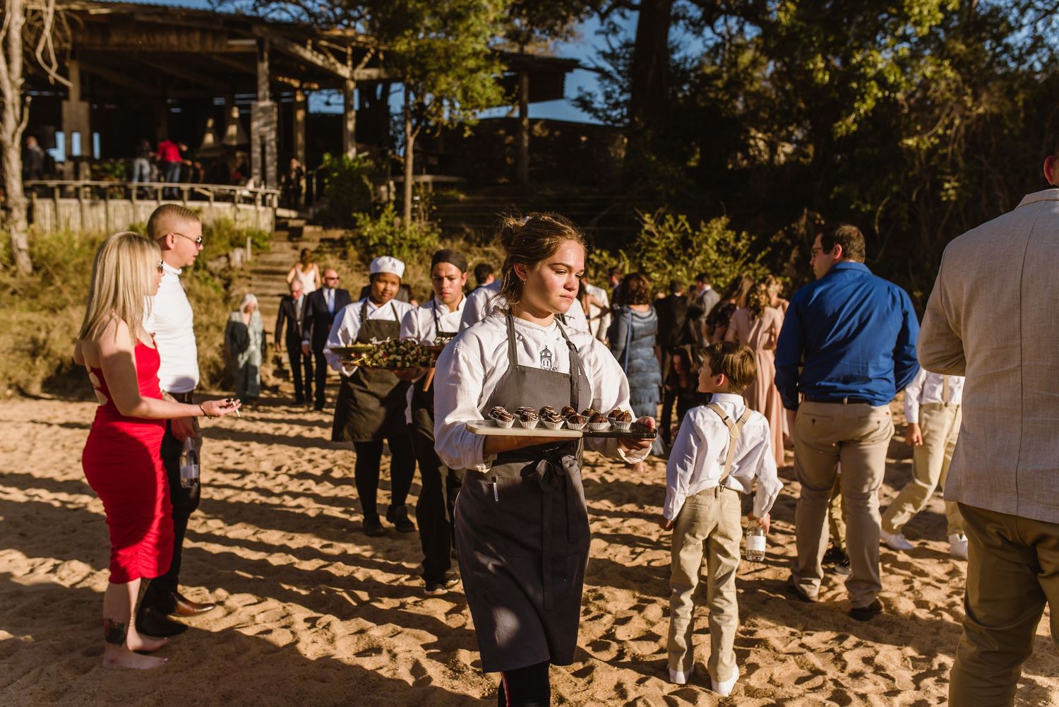 Guests gather at a beach wedding reception with trees and natural lighting in the background.