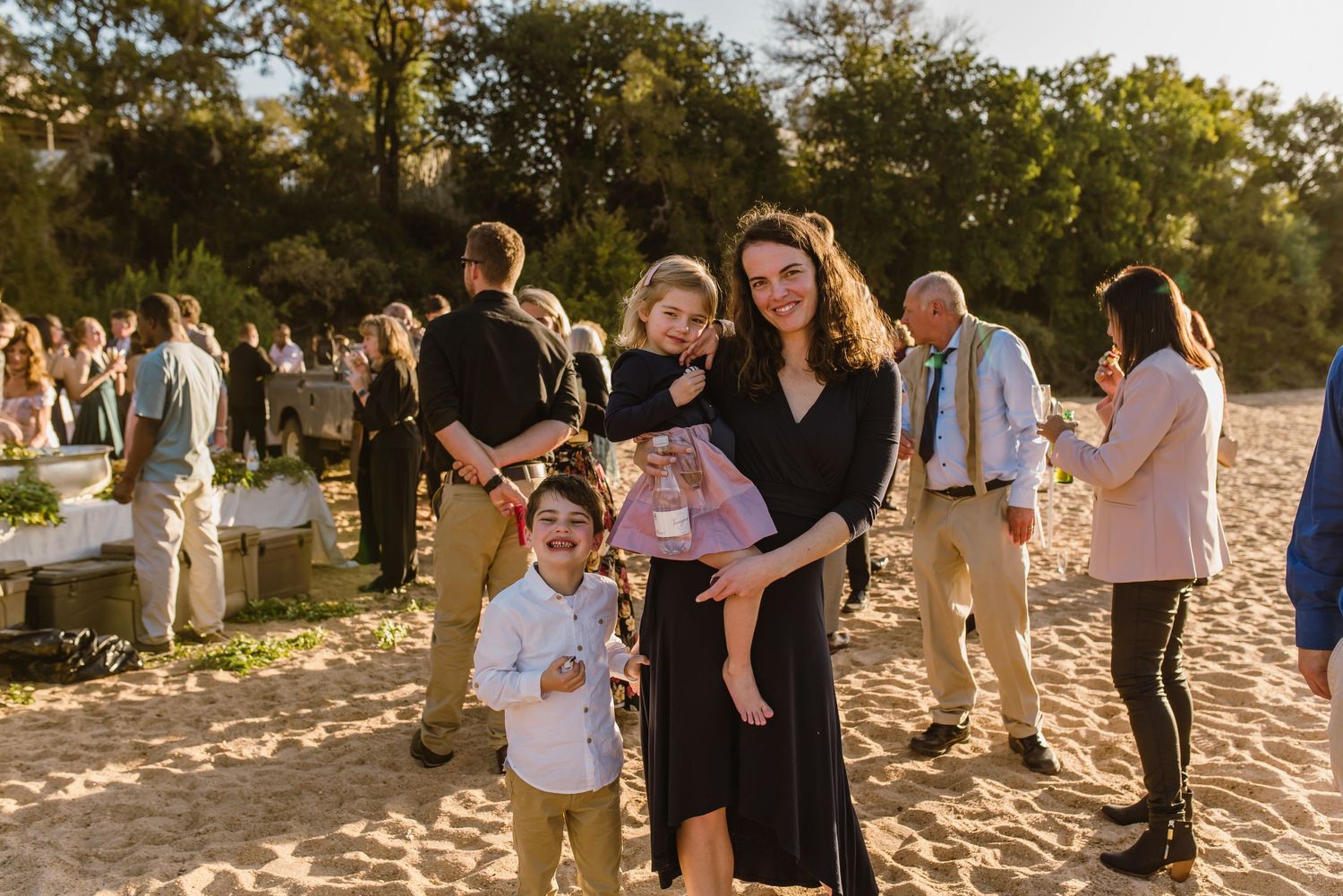 Wedding reception guests gather on a sandy beach surrounded by trees at sunset.