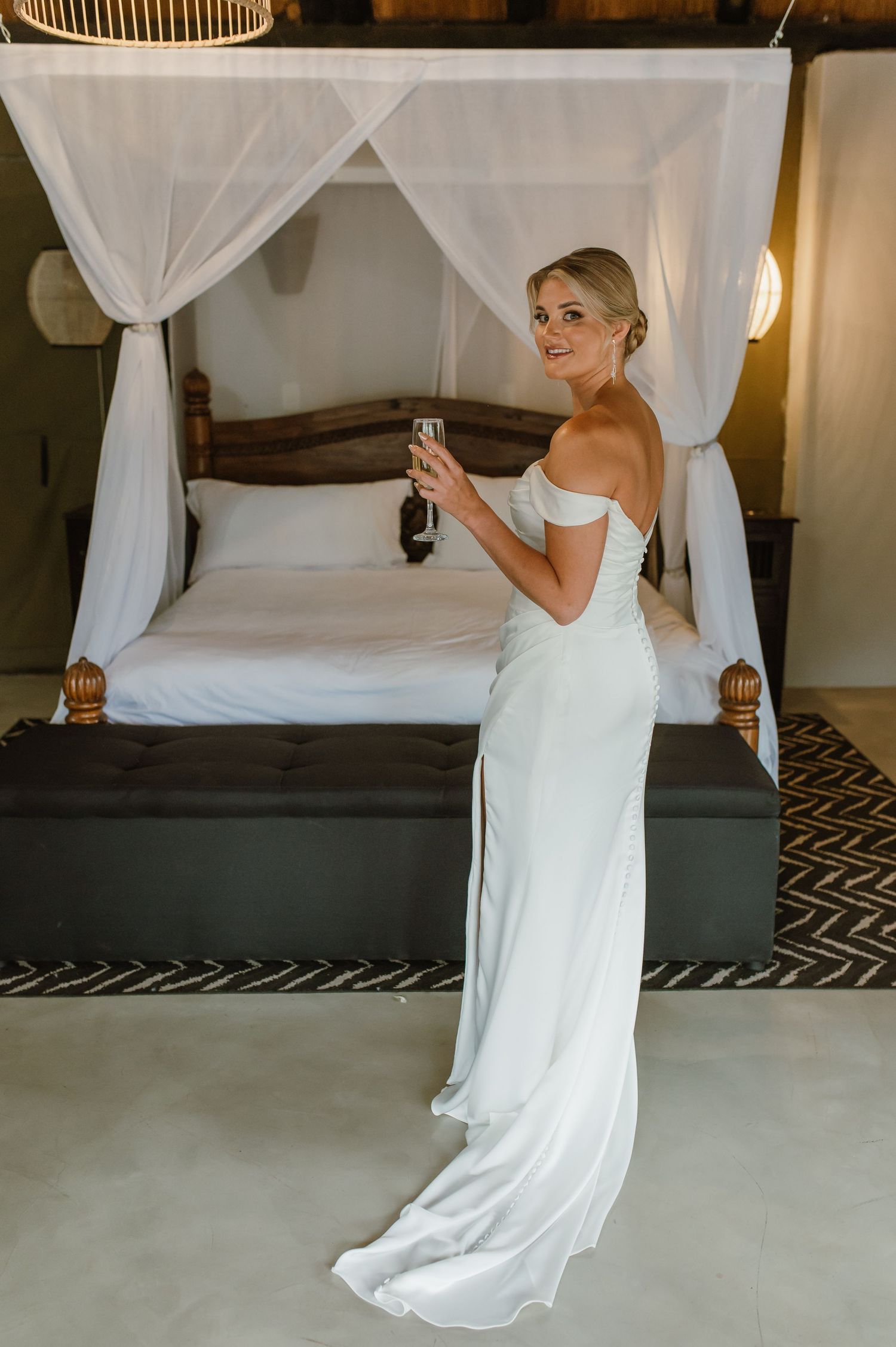 Elegant bride in white off-shoulder gown poses with champagne glass in front of canopy bed.