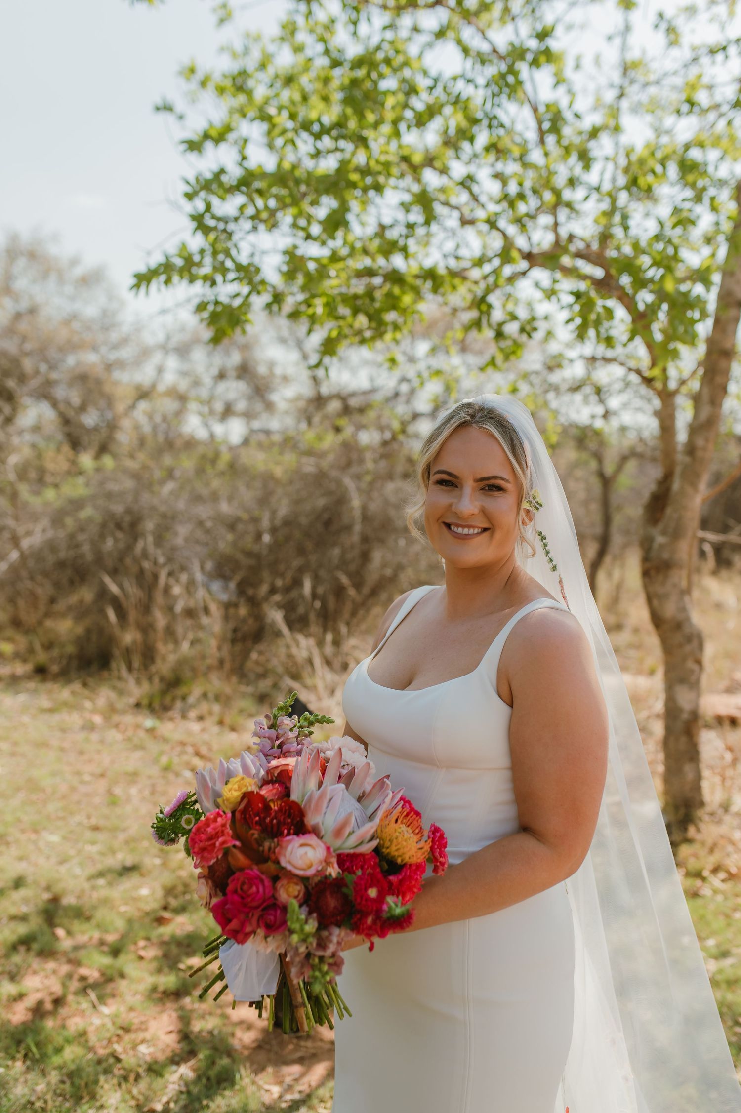 Bride holding vibrant red and pink bouquet against rustic outdoor backdrop.