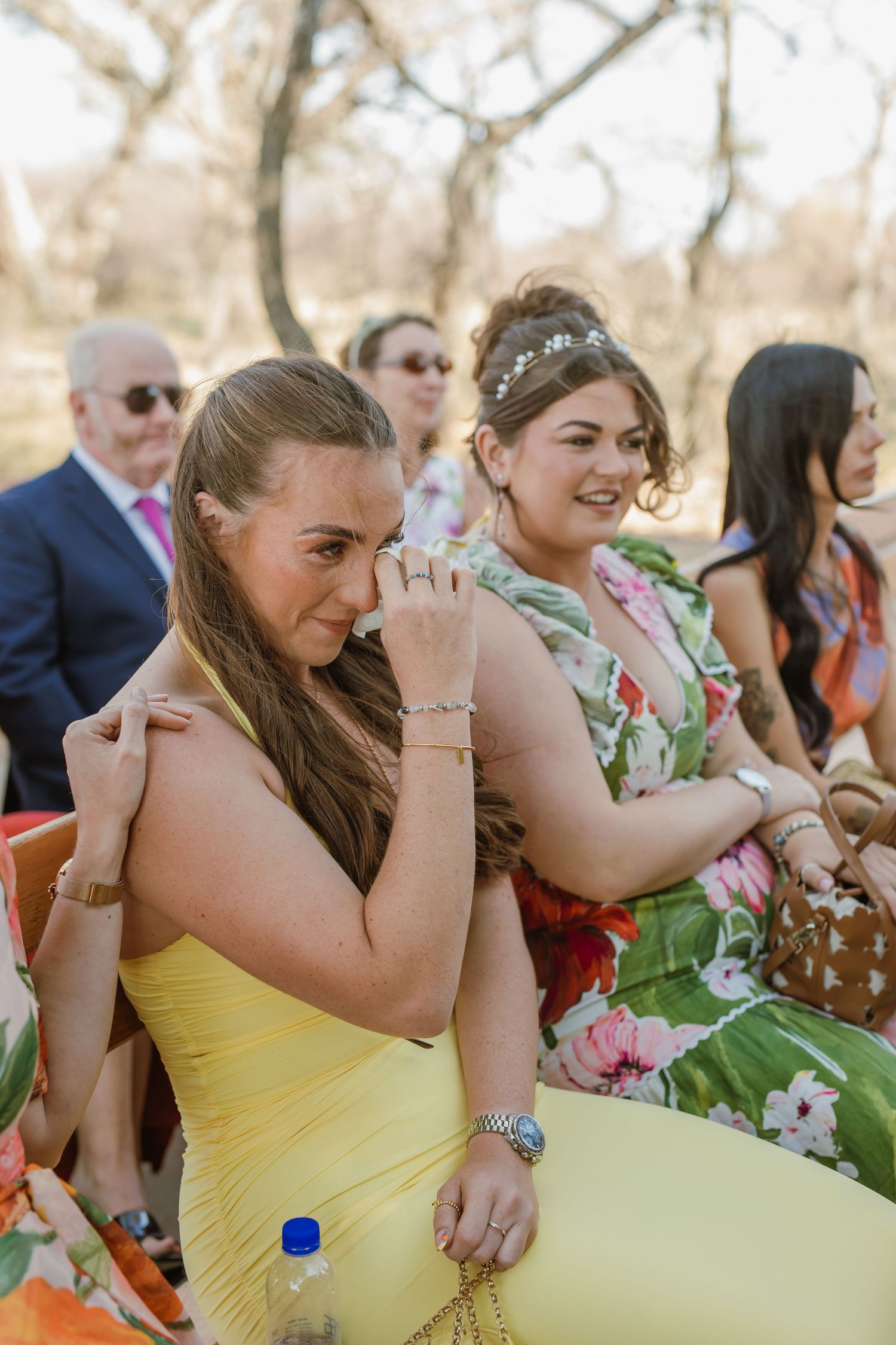 Bridesmaids in yellow dresses share emotional moments during an outdoor wedding ceremony.