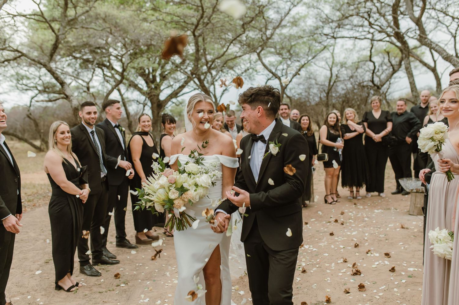 Newlyweds walk through confetti shower with wedding party in black attire.