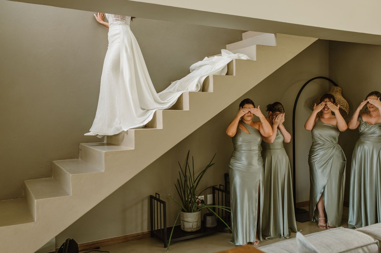 Wedding party in sage green dresses poses together on an elegant white staircase.