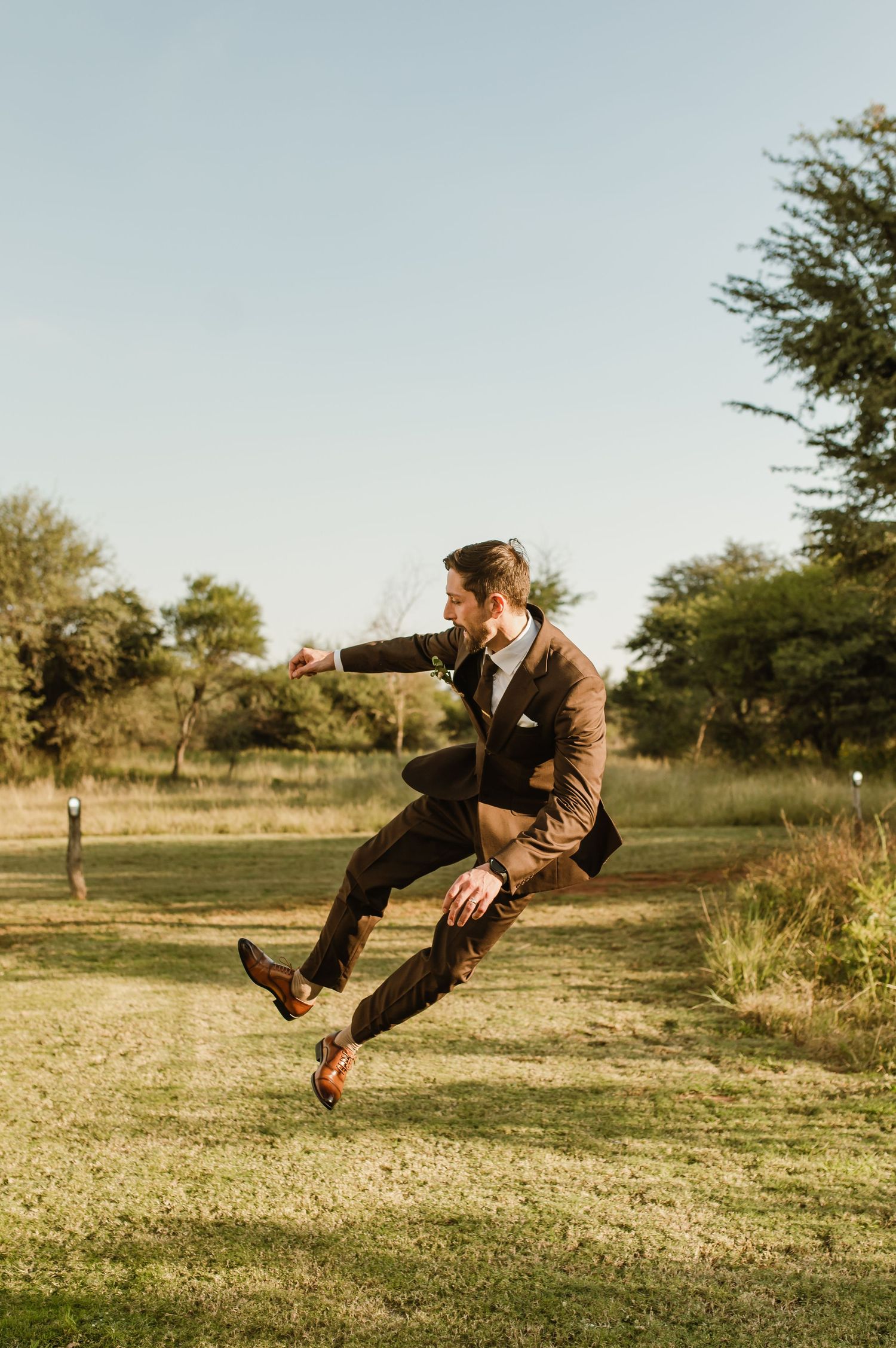 Groom in brown suit performs dramatic jump pose outdoors in natural setting.