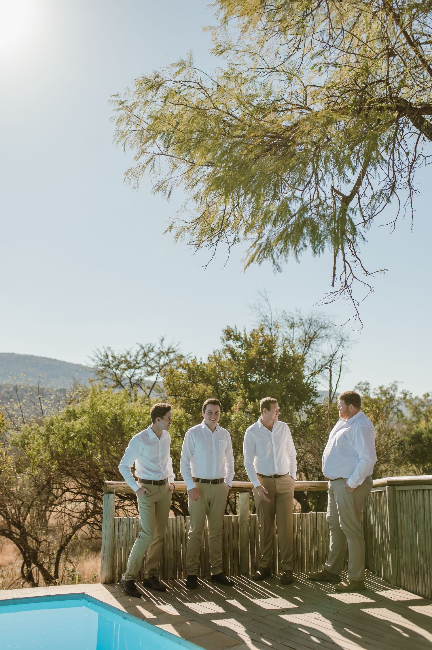 Four men in light colored clothing posing on deck with pool and mountain view.
