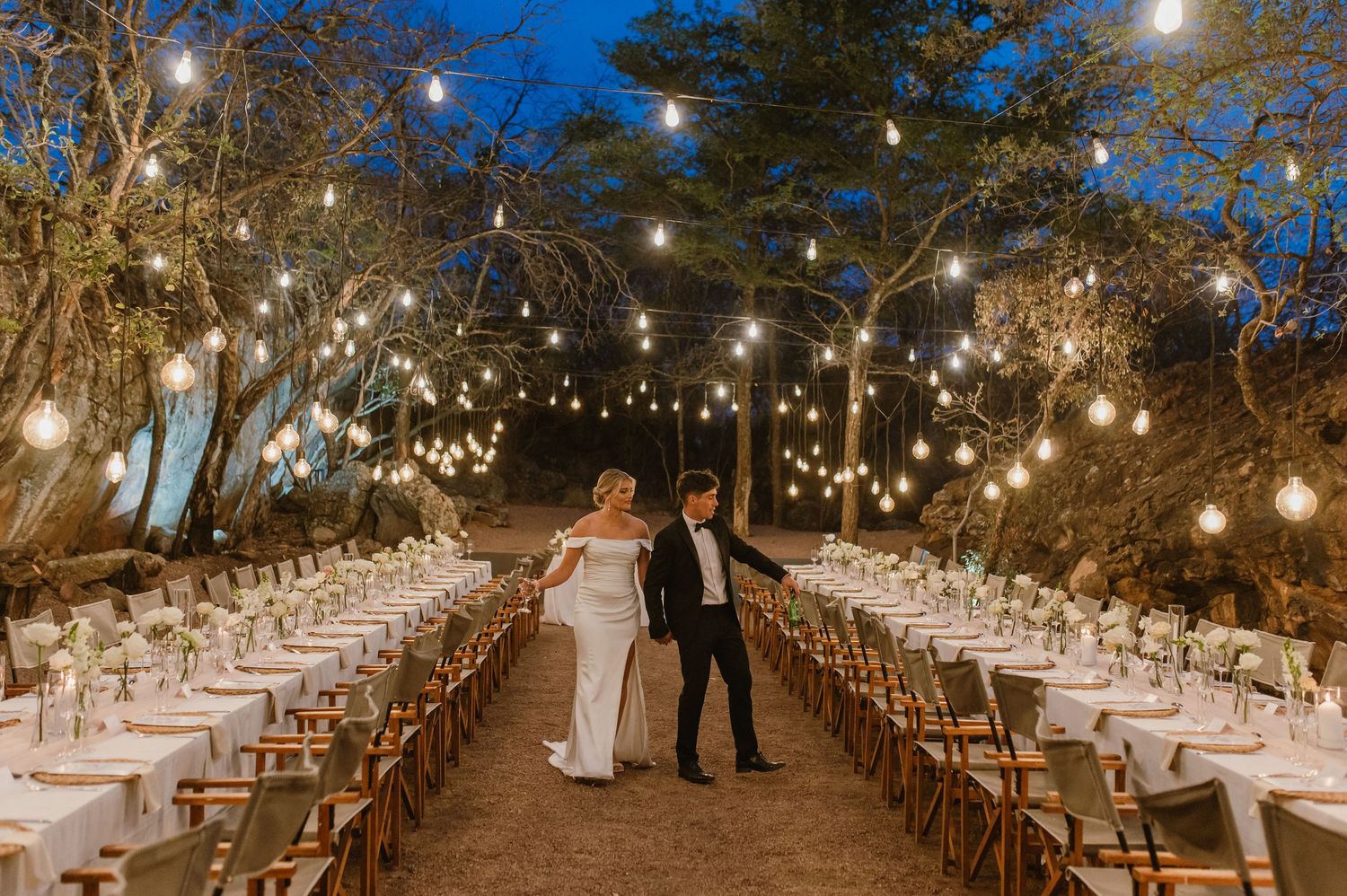 Rustic outdoor wedding venue with long dining tables illuminated by overhead string lights at twilight.