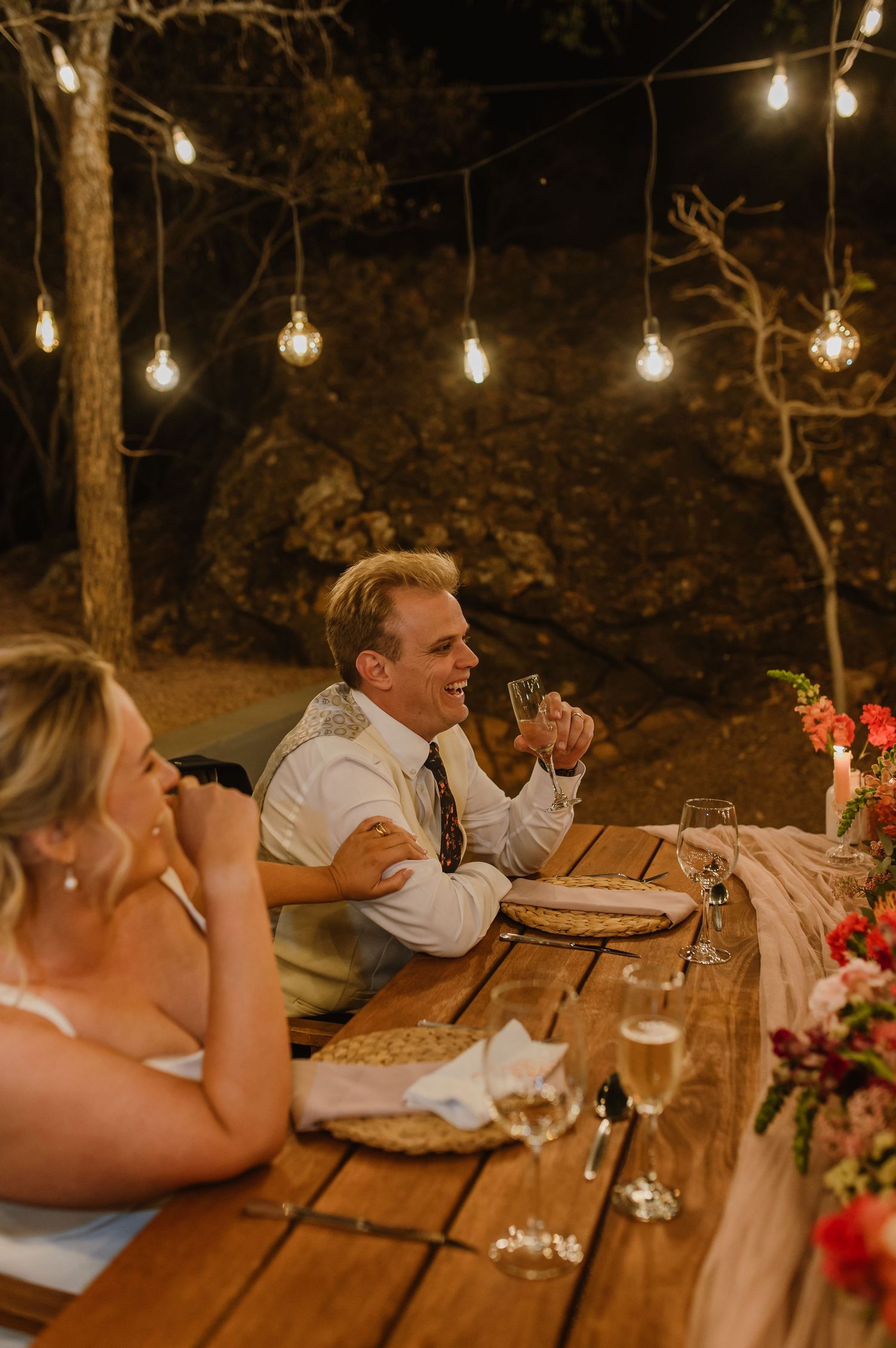 Diners seated at a long wooden table beneath twinkling lights in a natural outdoor setting.