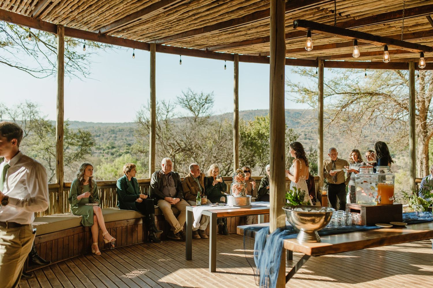 Guests gather at a rustic outdoor venue with wooden roof and scenic desert views in the background.