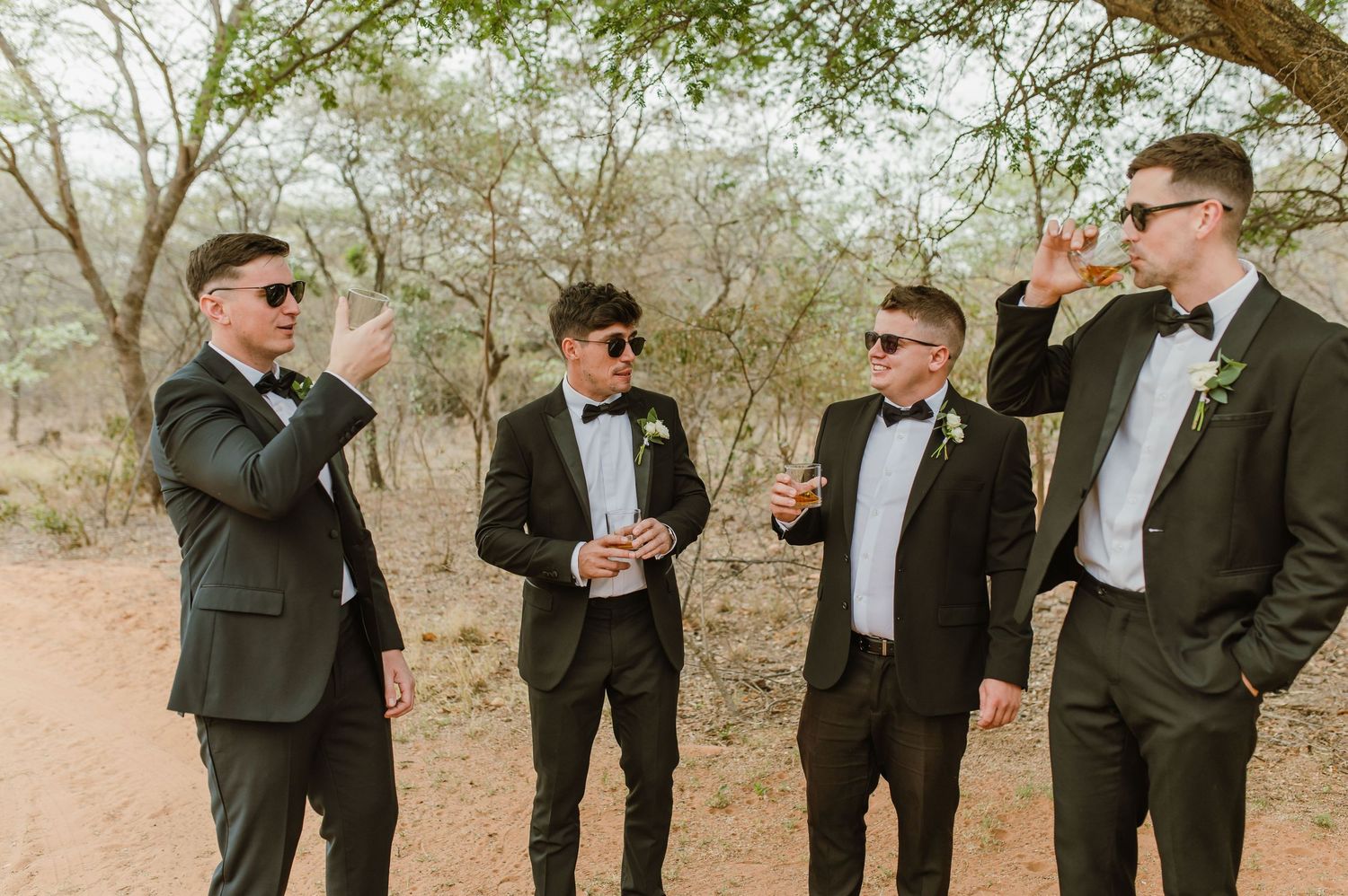 Groomsmen in black tuxedos and bow ties posing casually outdoors among trees.