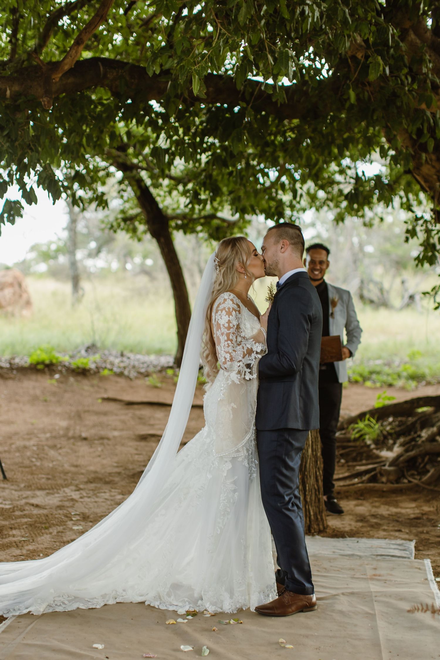 A romantic first kiss moment during an outdoor wedding ceremony beneath leafy trees.