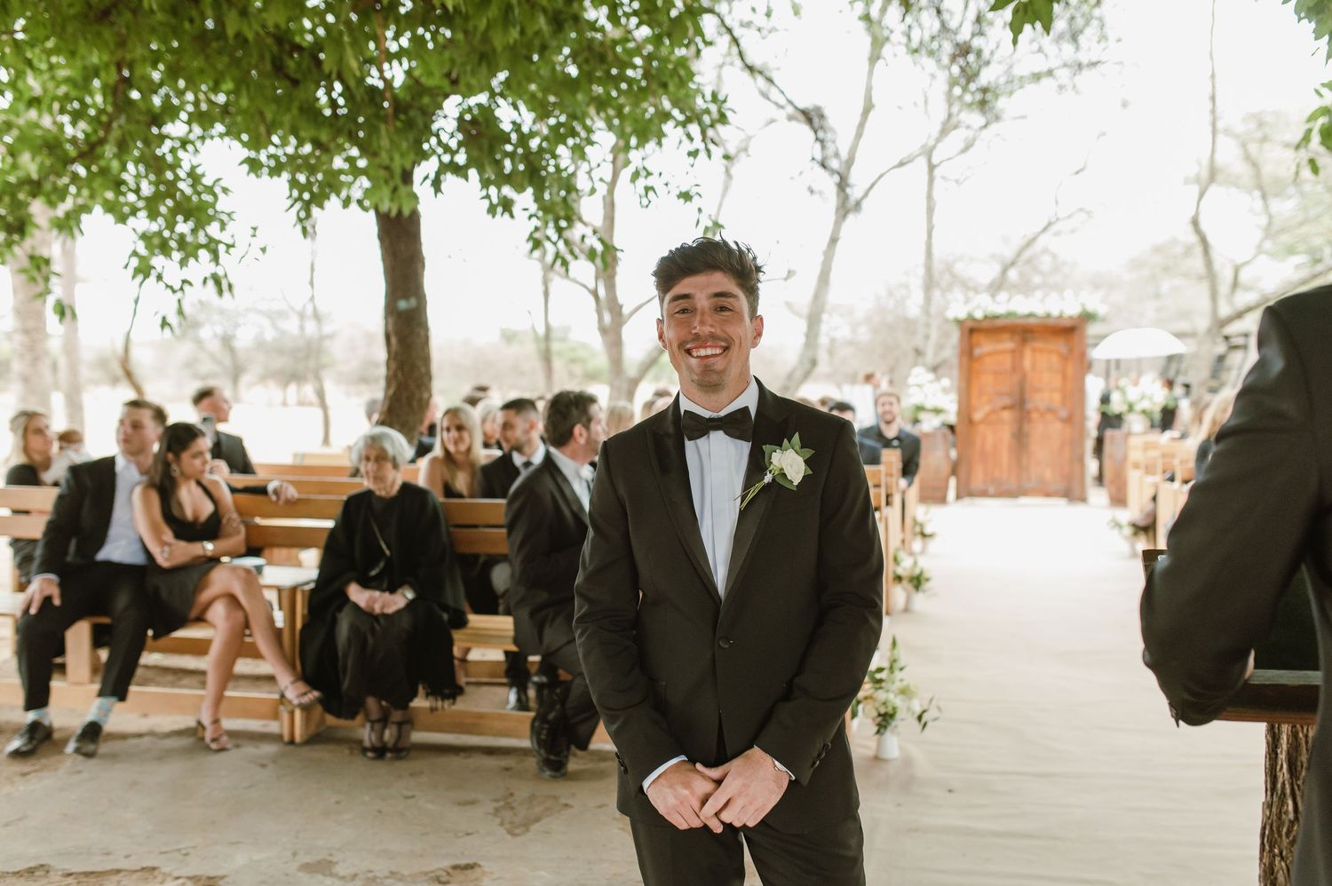 Formal wedding party members stand beneath green leafy trees during outdoor celebration.