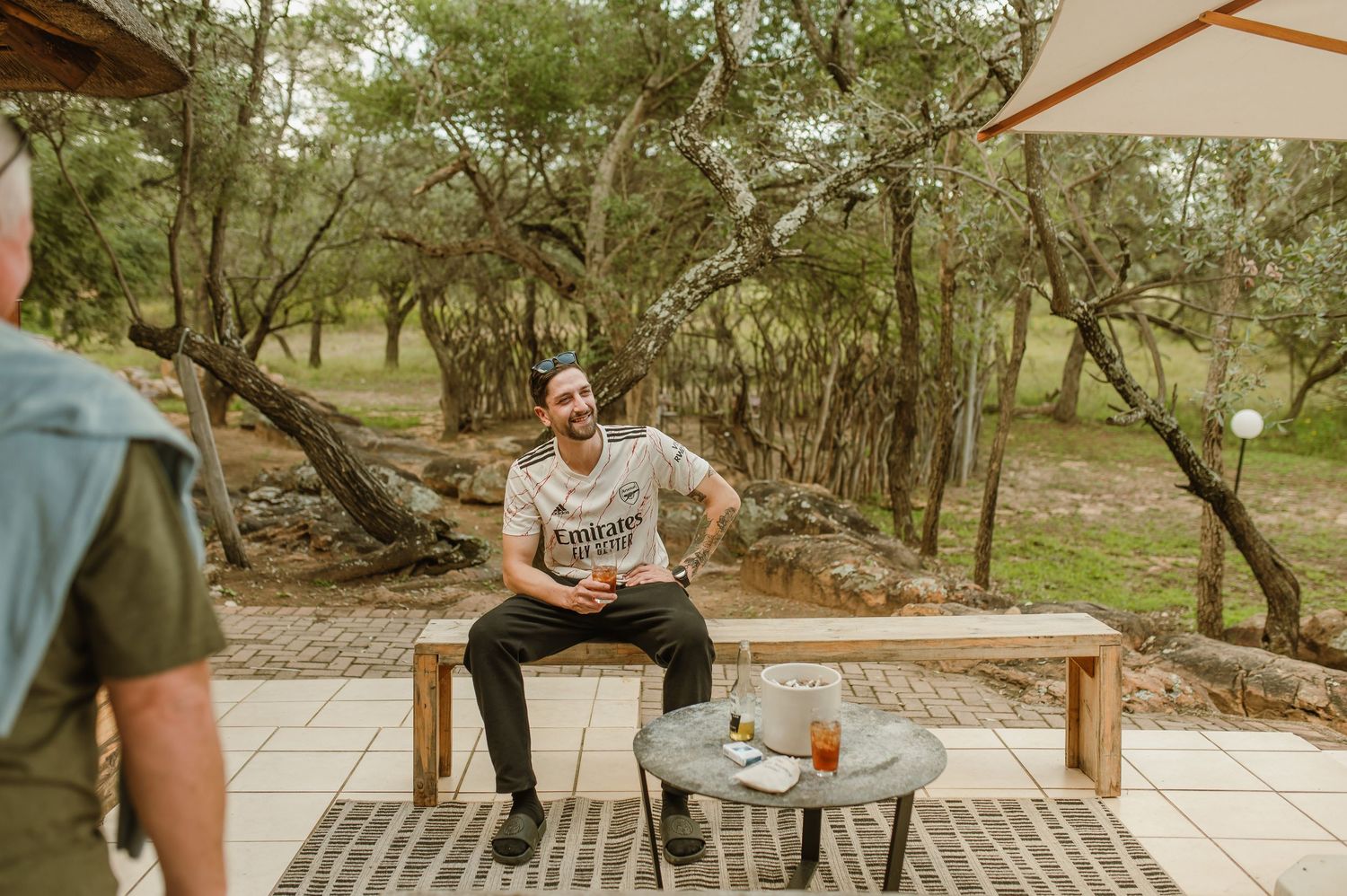 People enjoying conversation and drinks at a covered outdoor seating area surrounded by trees.