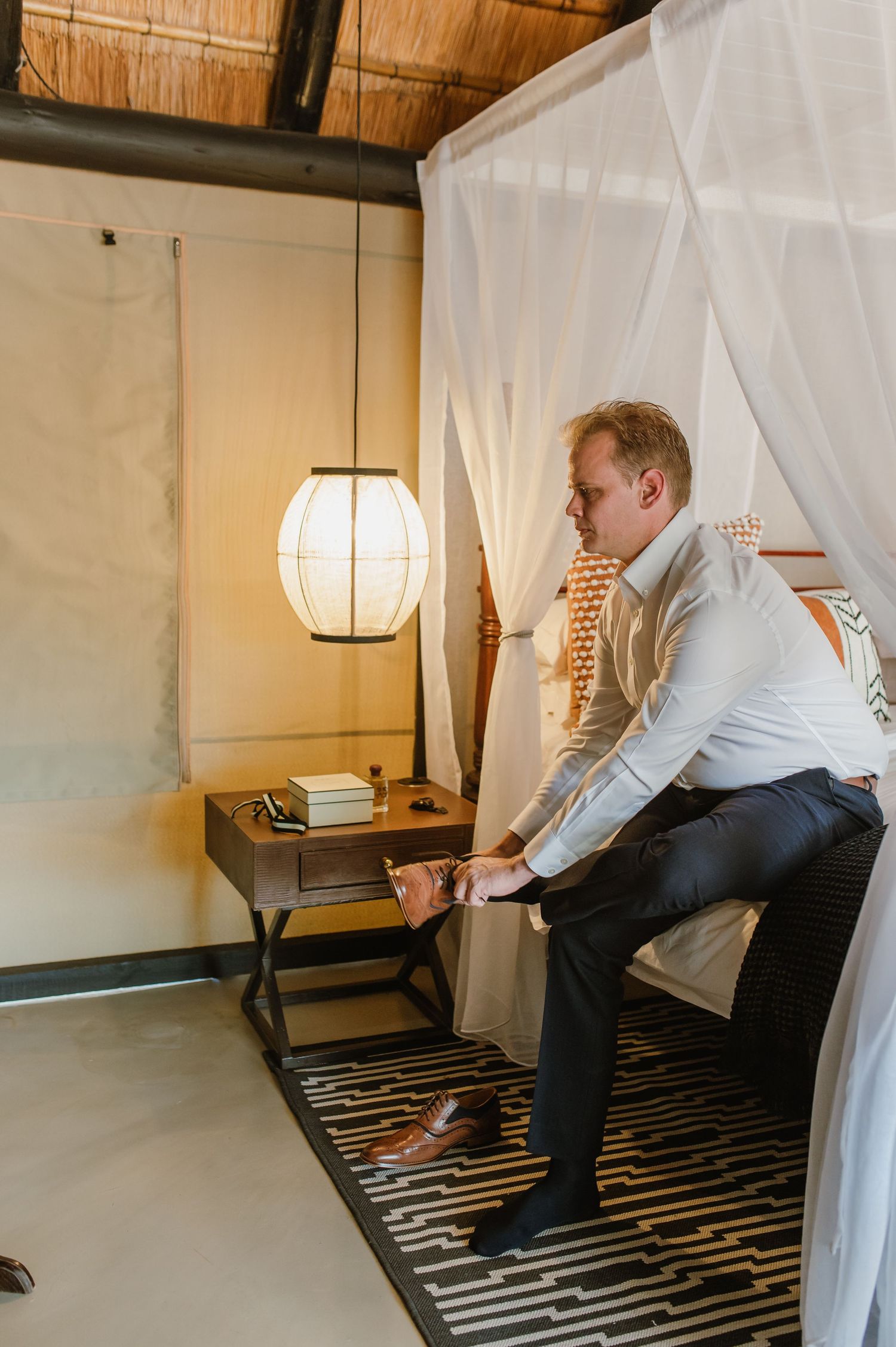Person in formal attire adjusts items on a bedside table in a luxurious room with sheer curtains.