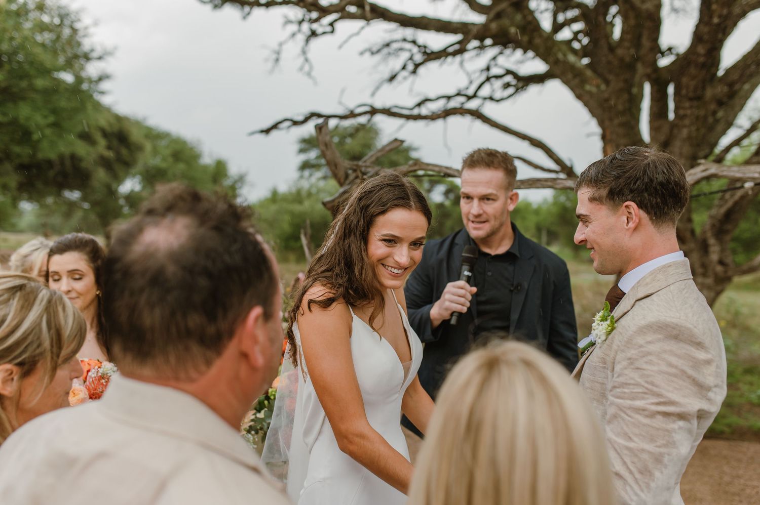 Wedding guests gather around a white ceremony space under a large tree with floral decorations.