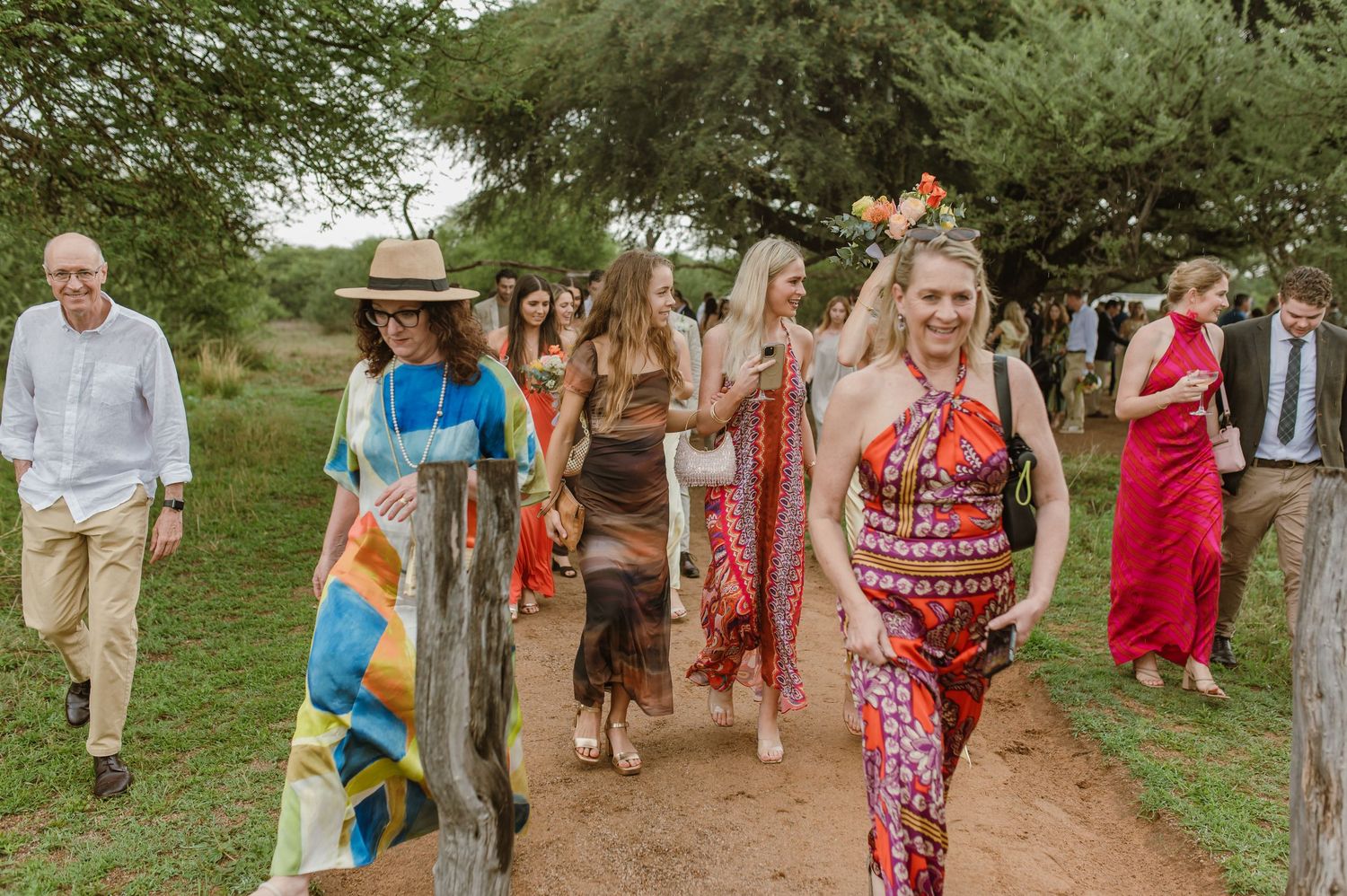 Colorfully dressed wedding guests walking along a dirt path under trees.