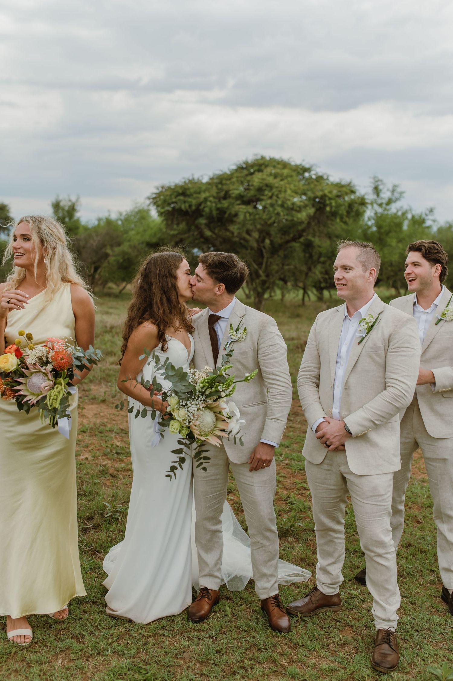 Wedding party laughing and interacting in casual poses on grassy field.