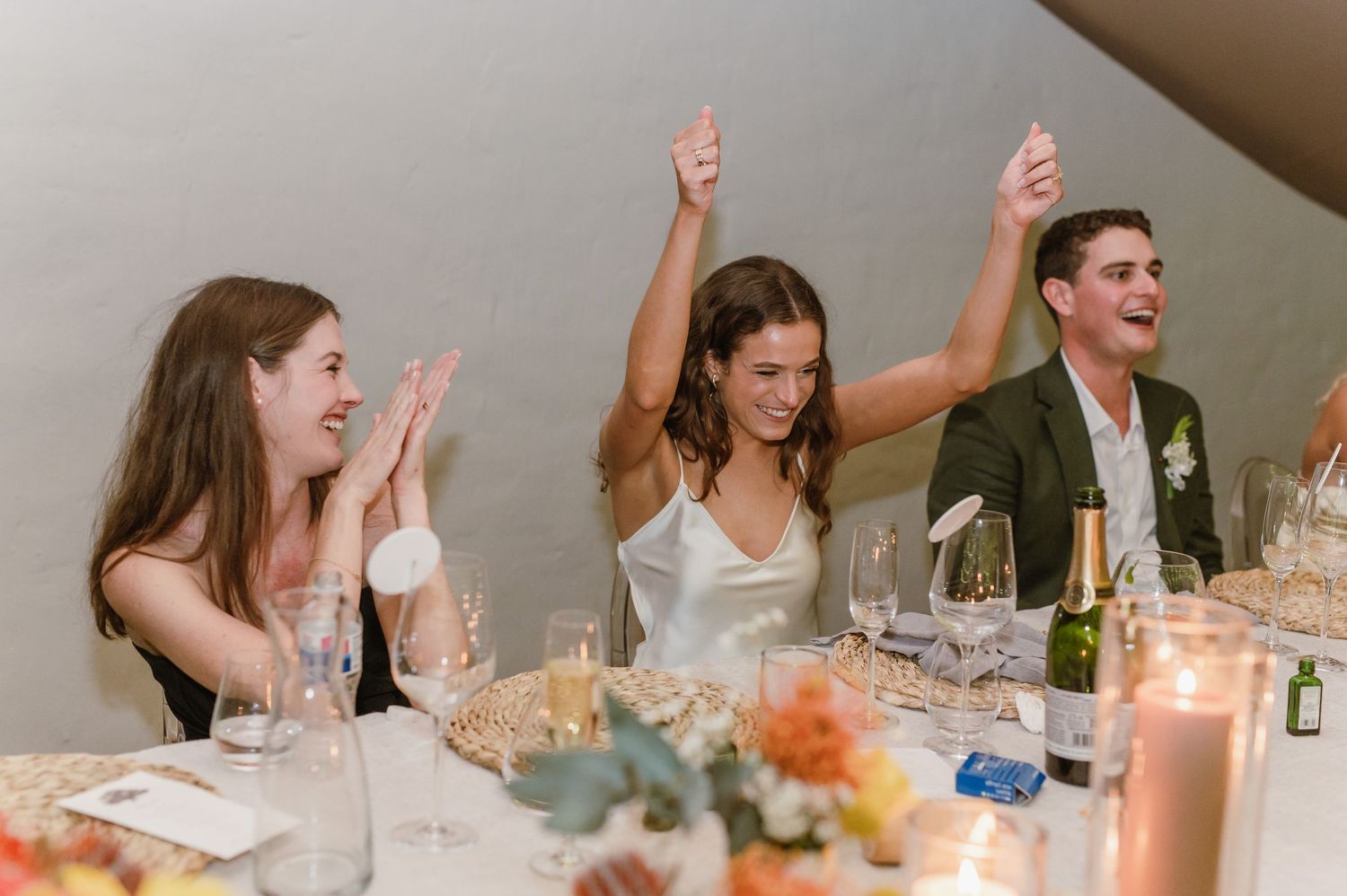 Wedding guests cheering and raising their hands during reception celebrations at decorated tables.