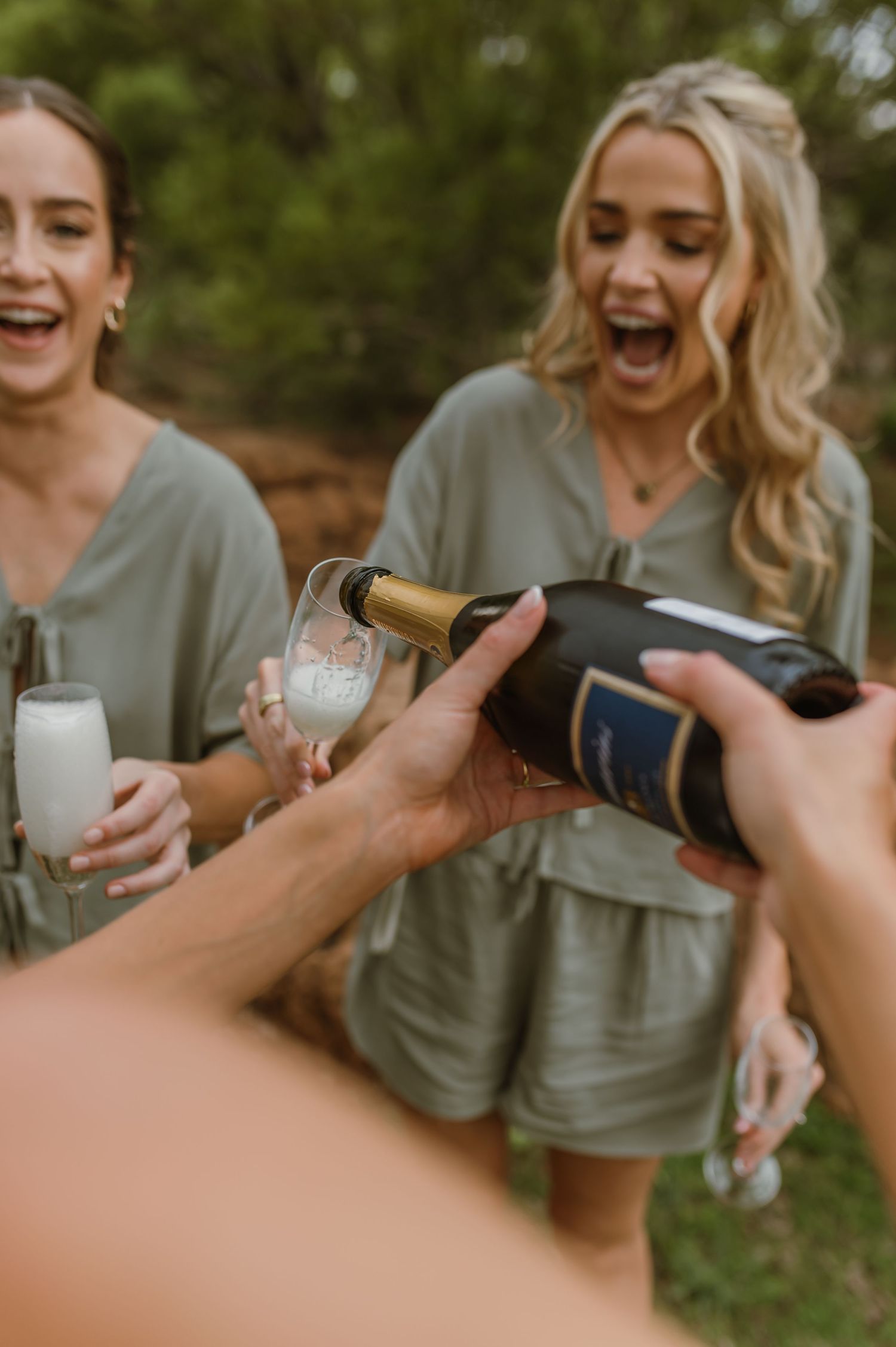 Friends in matching sage rompers laugh joyfully while champagne is being poured into glasses outdoors.