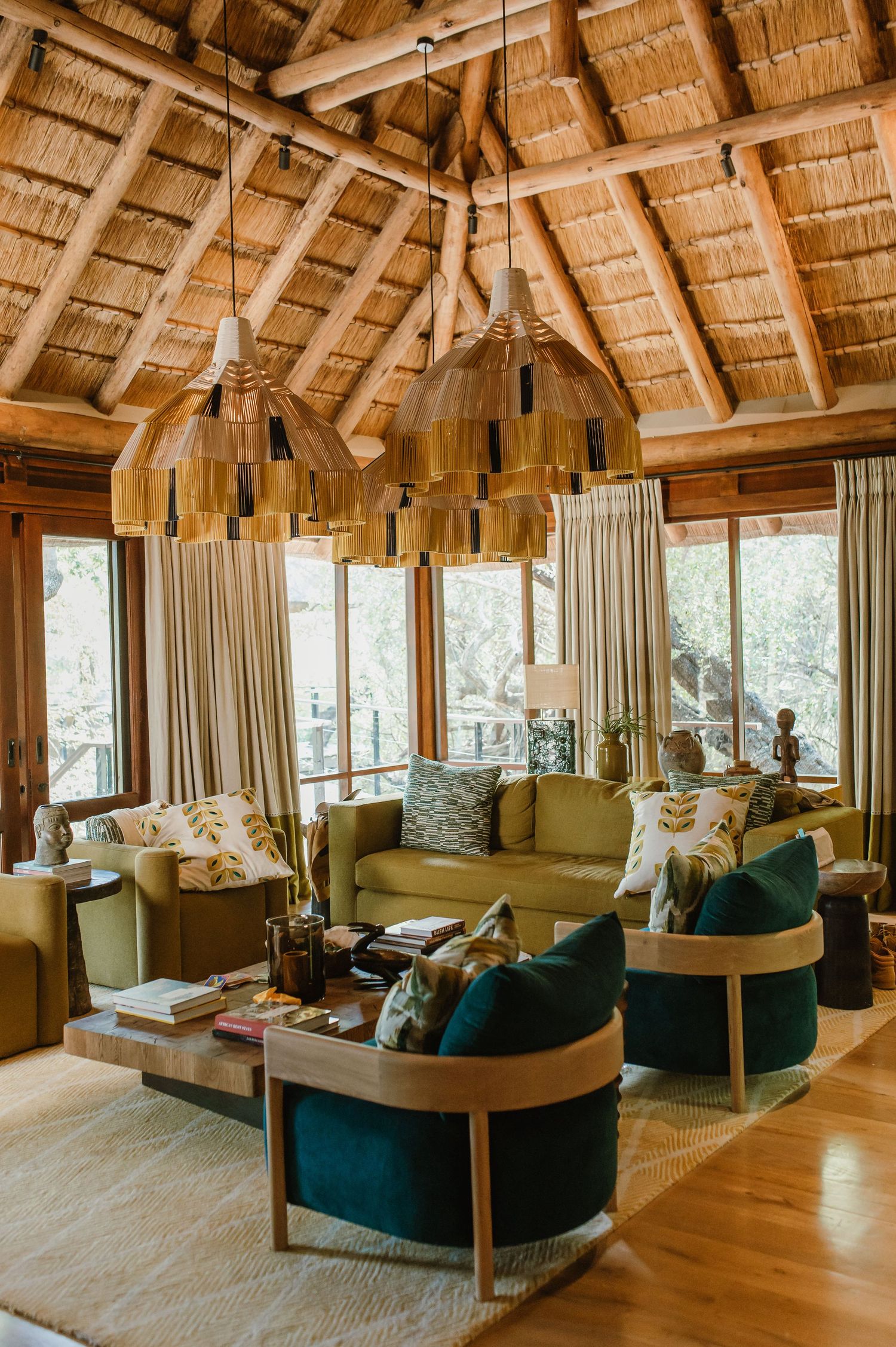 Luxury safari lodge living room with thatched roof, pendant lights, green velvet chairs, and olive sofas.
