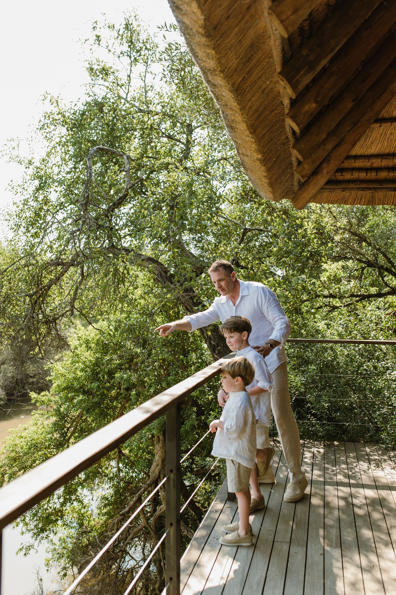 Father pointing at nature with two young boys on wooden deck overlooking lush greenery.